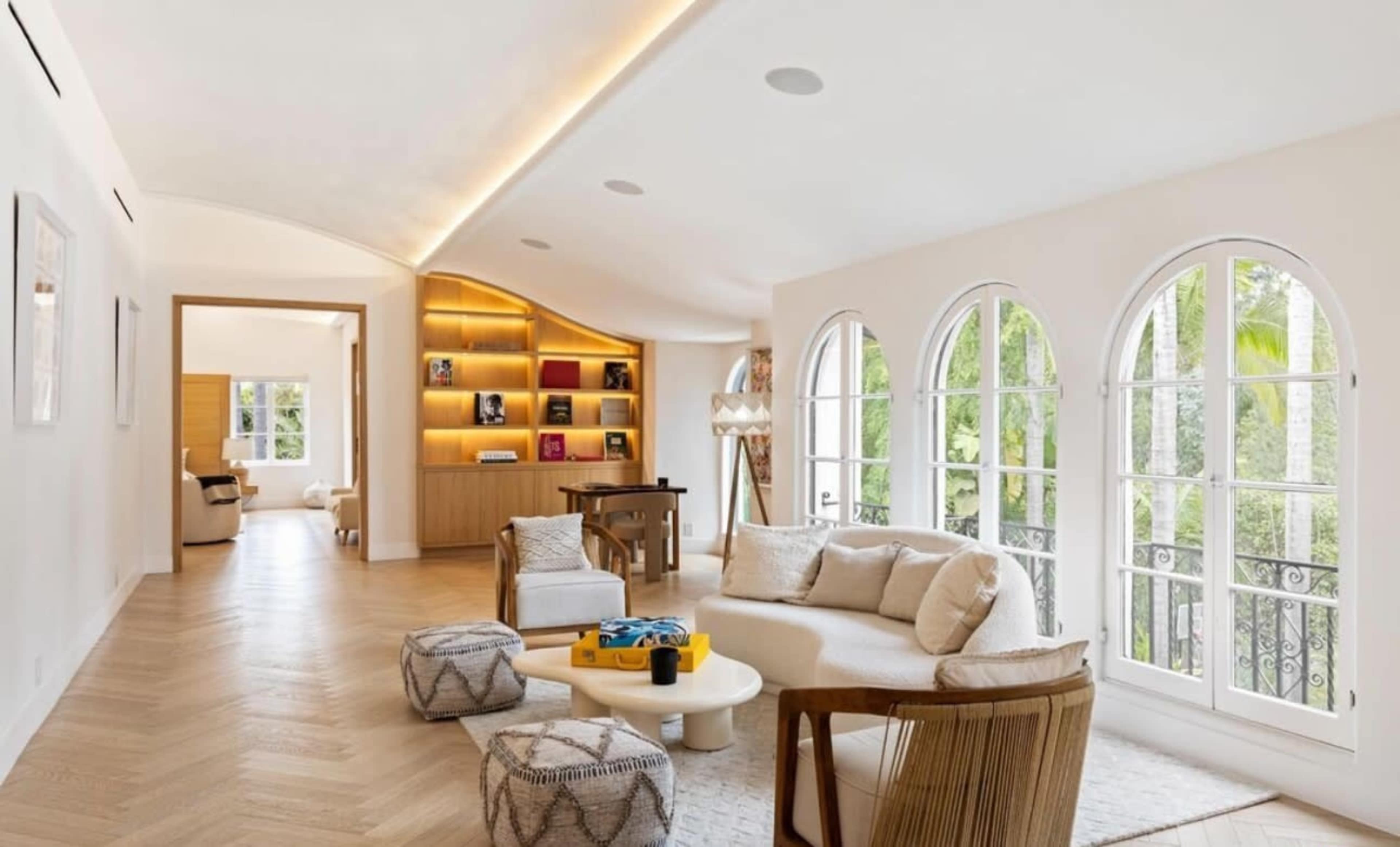 The image shows a brightly lit, elegantly designed living room with large arched windows, modern furniture, and a light wooden floor.