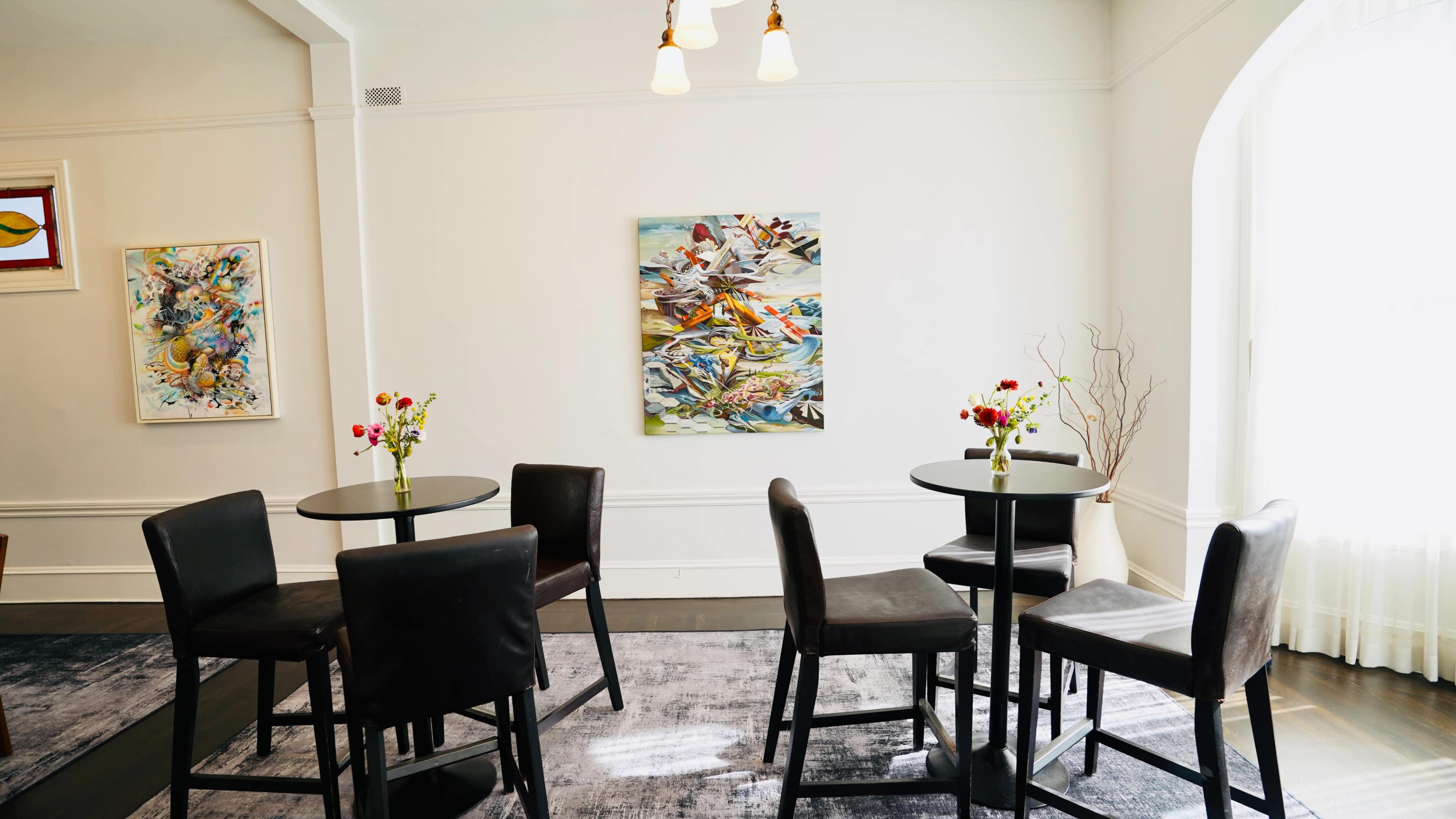 A brightly lit room with two small round tables, each adorned with a vase of flowers, and several black chairs arranged around them, alongside a colorful painting on the wall.