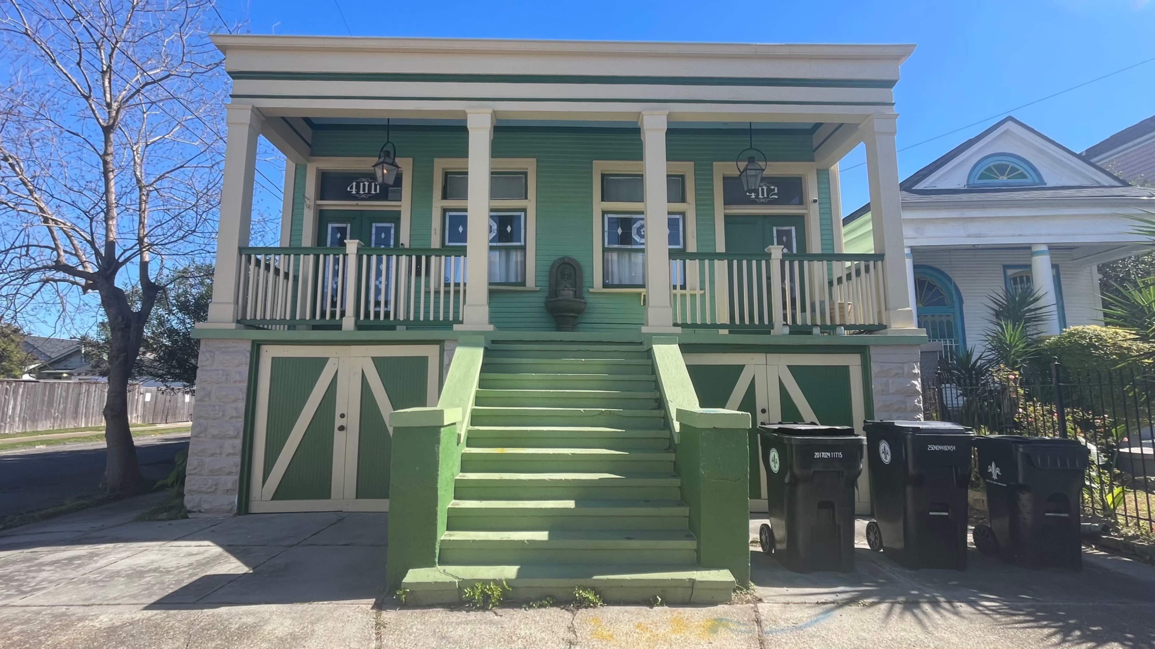 The image shows a two-story green house with white columns, a staircase leading up to two separate entrances, and three trash bins in front.