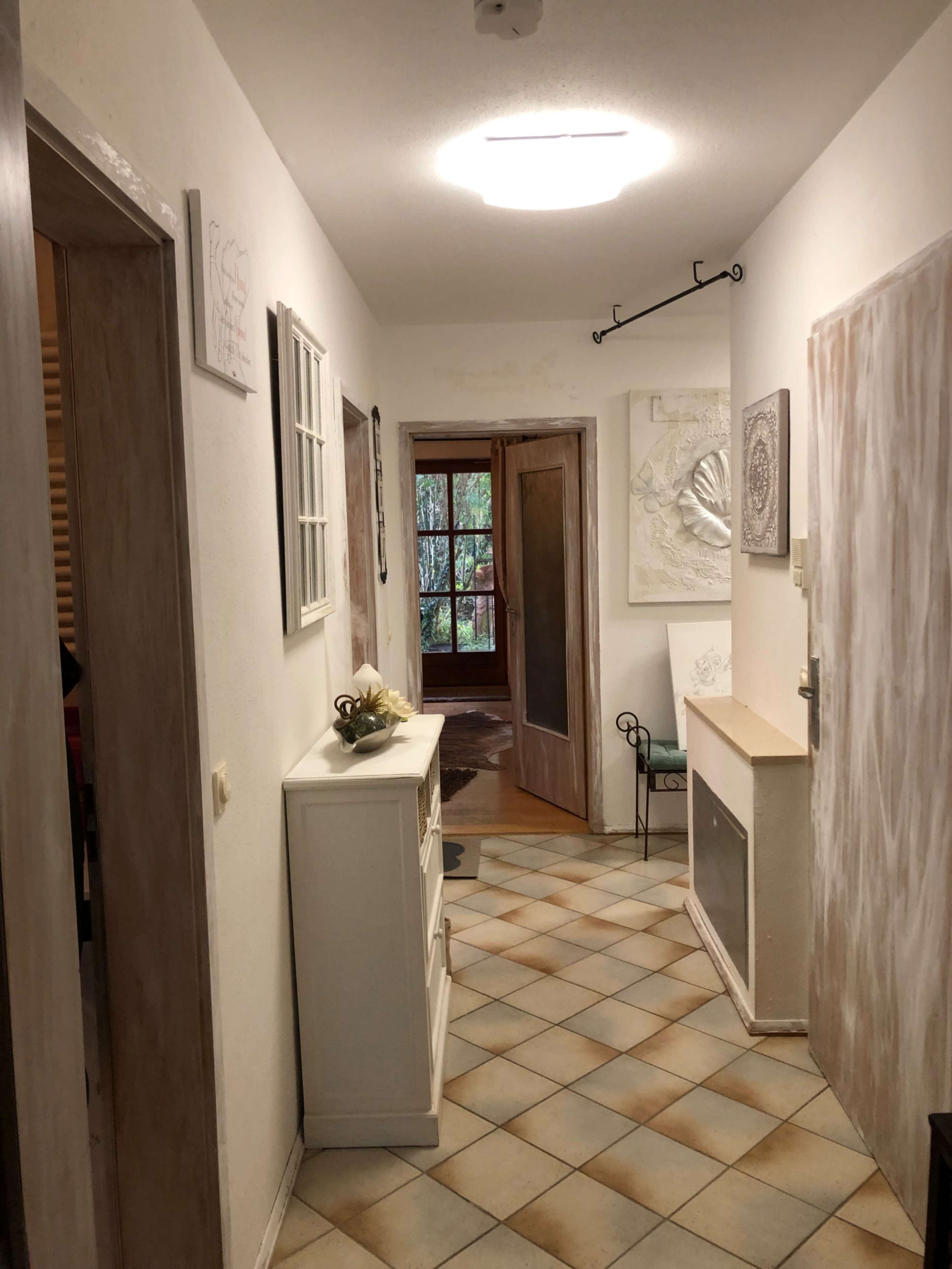 The image shows a hallway with tiled flooring, wooden doors, and a side table adorned with a decorative item, leading to an open door at the end.