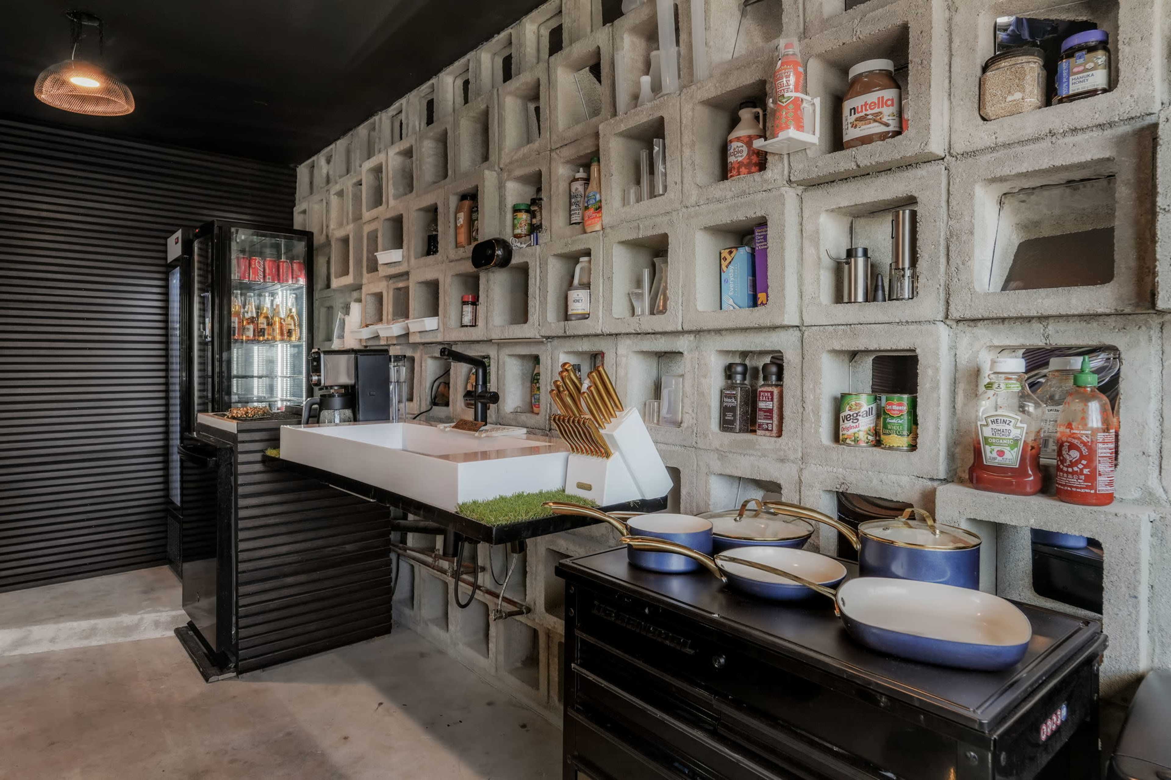 The image shows a modern kitchen featuring a concrete block wall with shelves displaying various condiments and kitchen items, alongside a sleek white sink and a dark cabinetry unit with cooking pots.