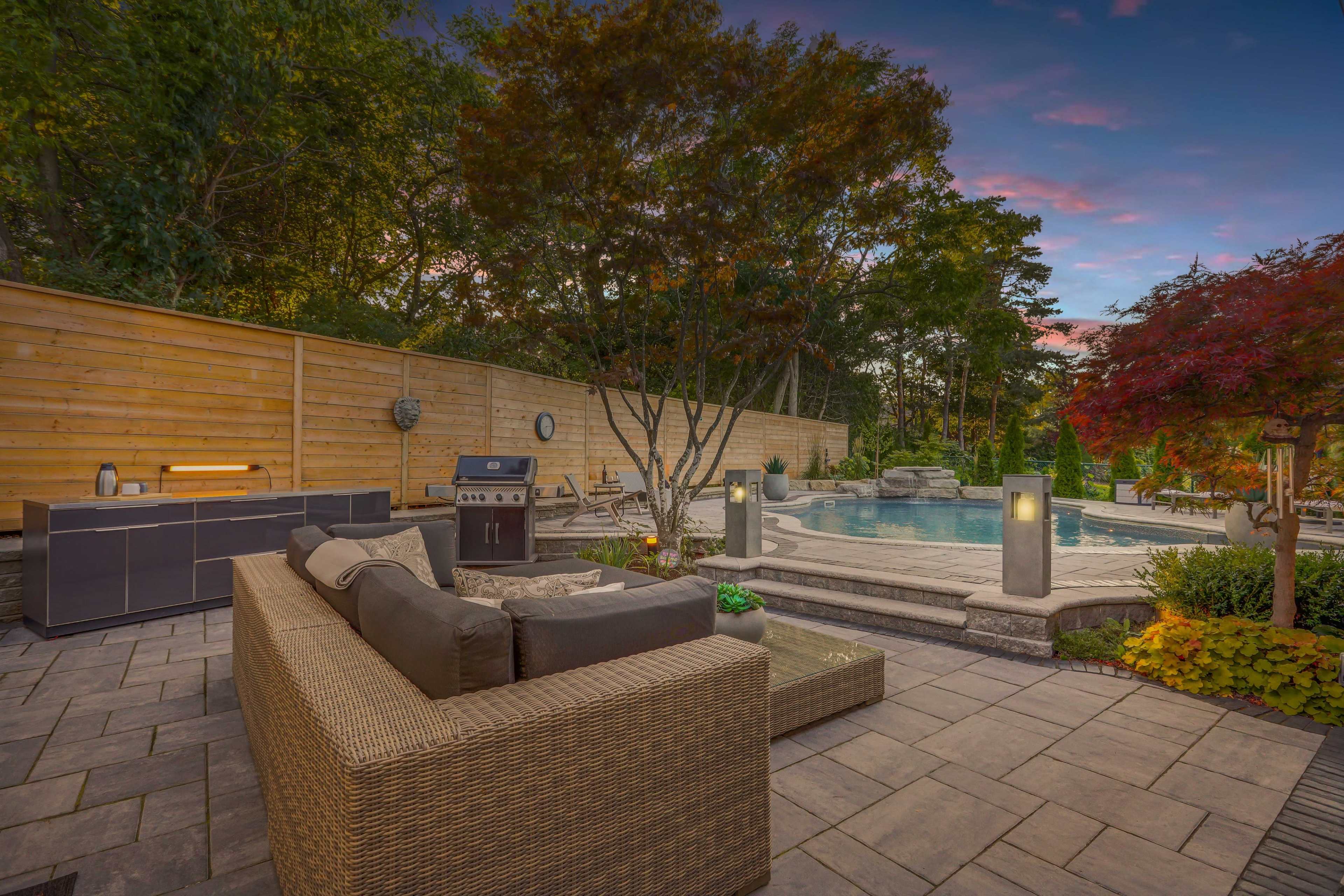 A backyard patio area with a stone path leading to a swimming pool, surrounded by trees and a wooden fence, featuring outdoor seating and a barbecue grill.