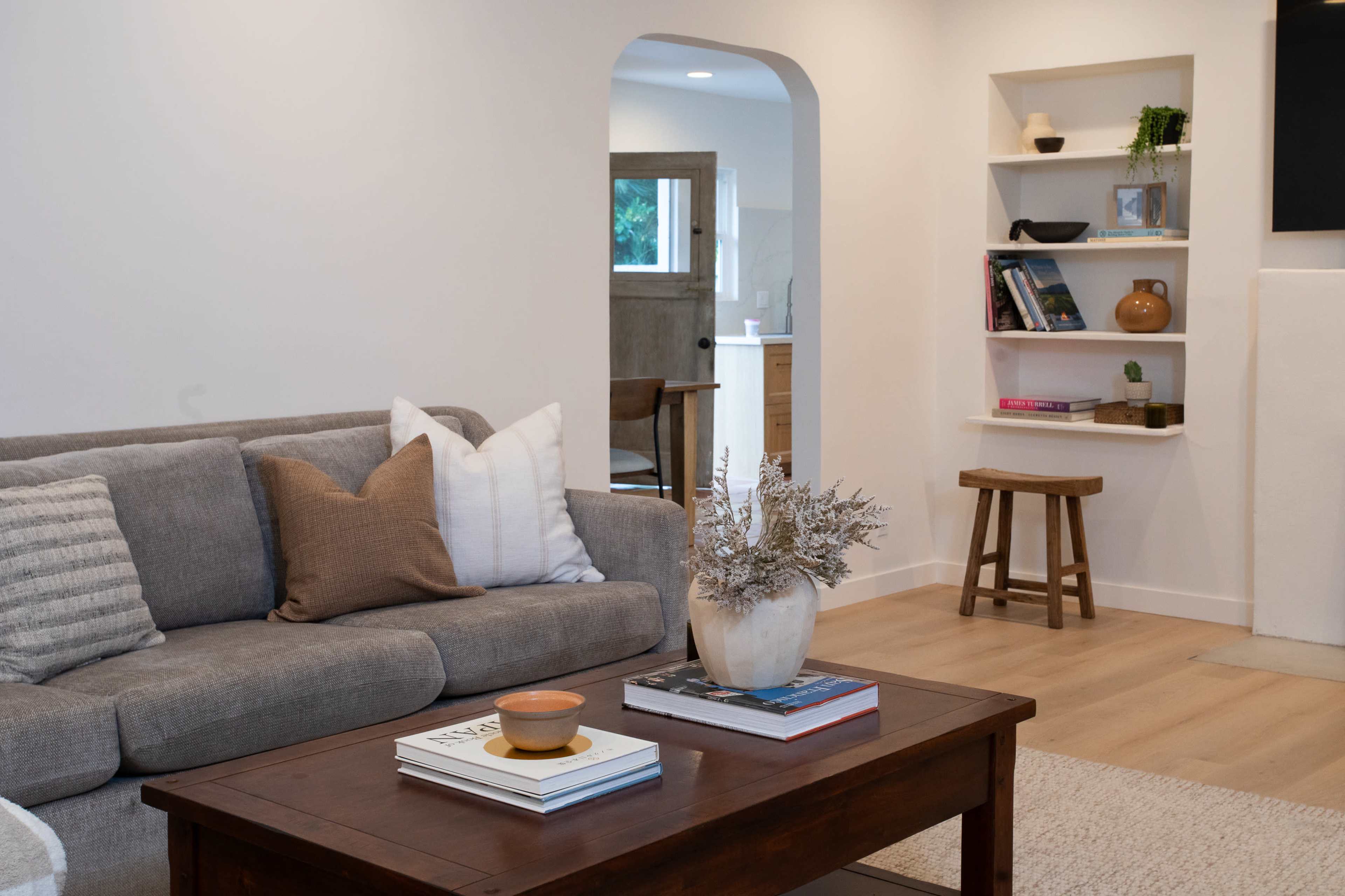 A light-colored living room features a grey sofa with decorative pillows, a wooden coffee table with books, and a small shelf displaying objects in the background.