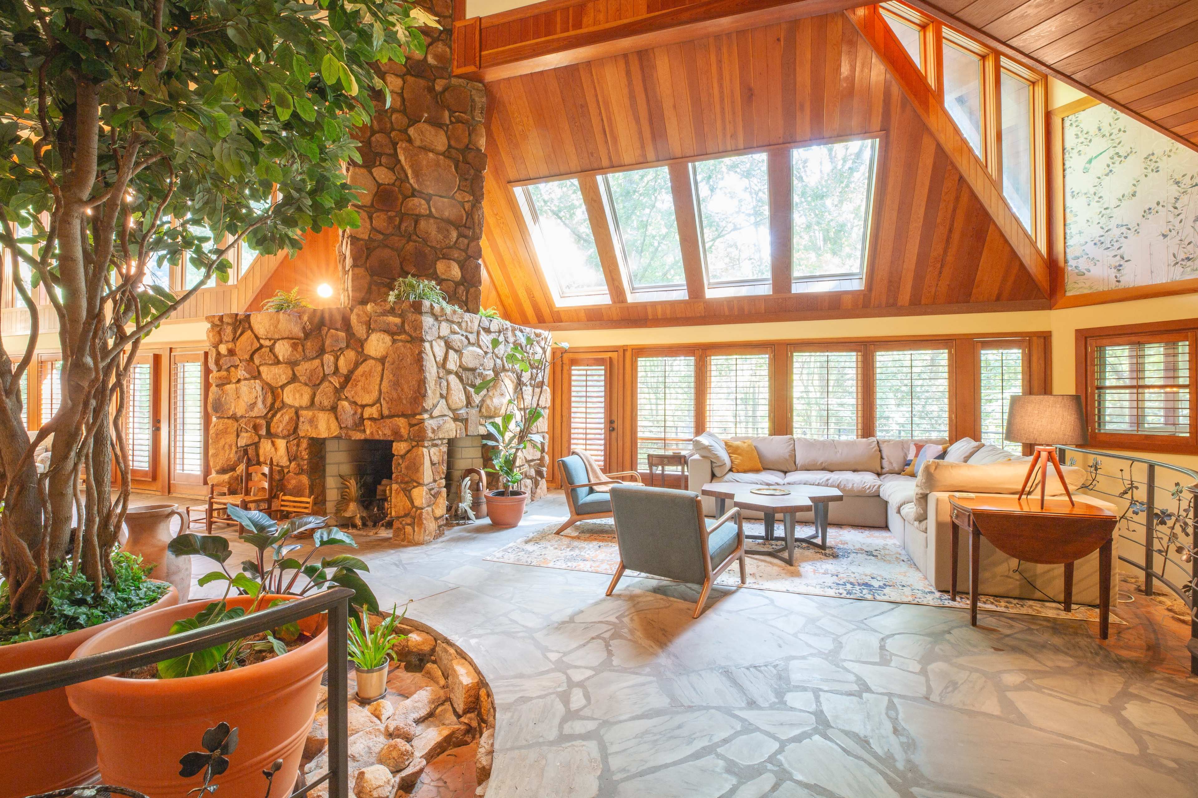 A spacious living room featuring a large stone fireplace, a seating area with a sofa and chairs, and large windows allowing ample natural light.