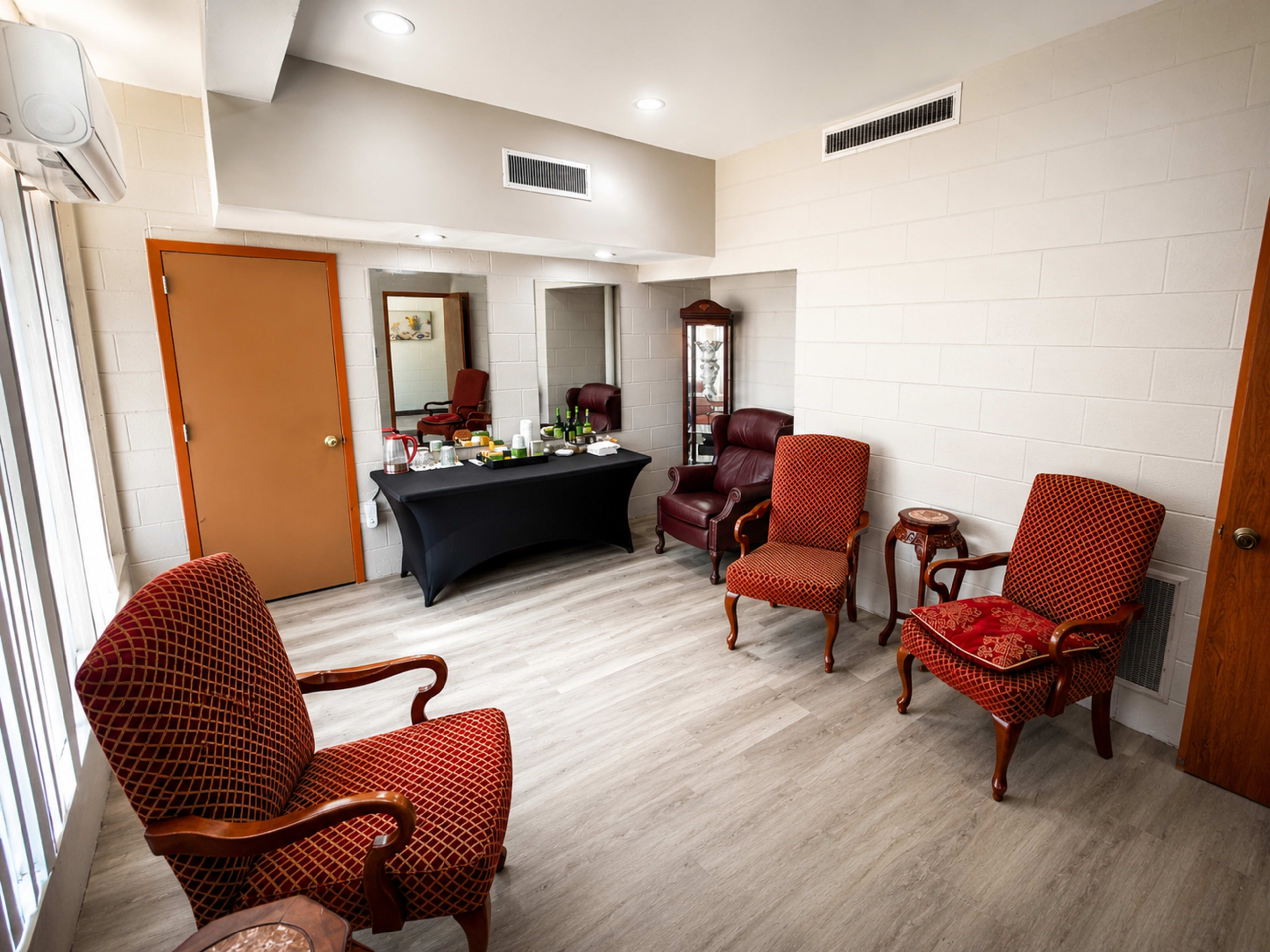 The image depicts a small room featuring two red upholstered chairs, a dark table with items on it, a full-length mirror, and a door leading to another area.