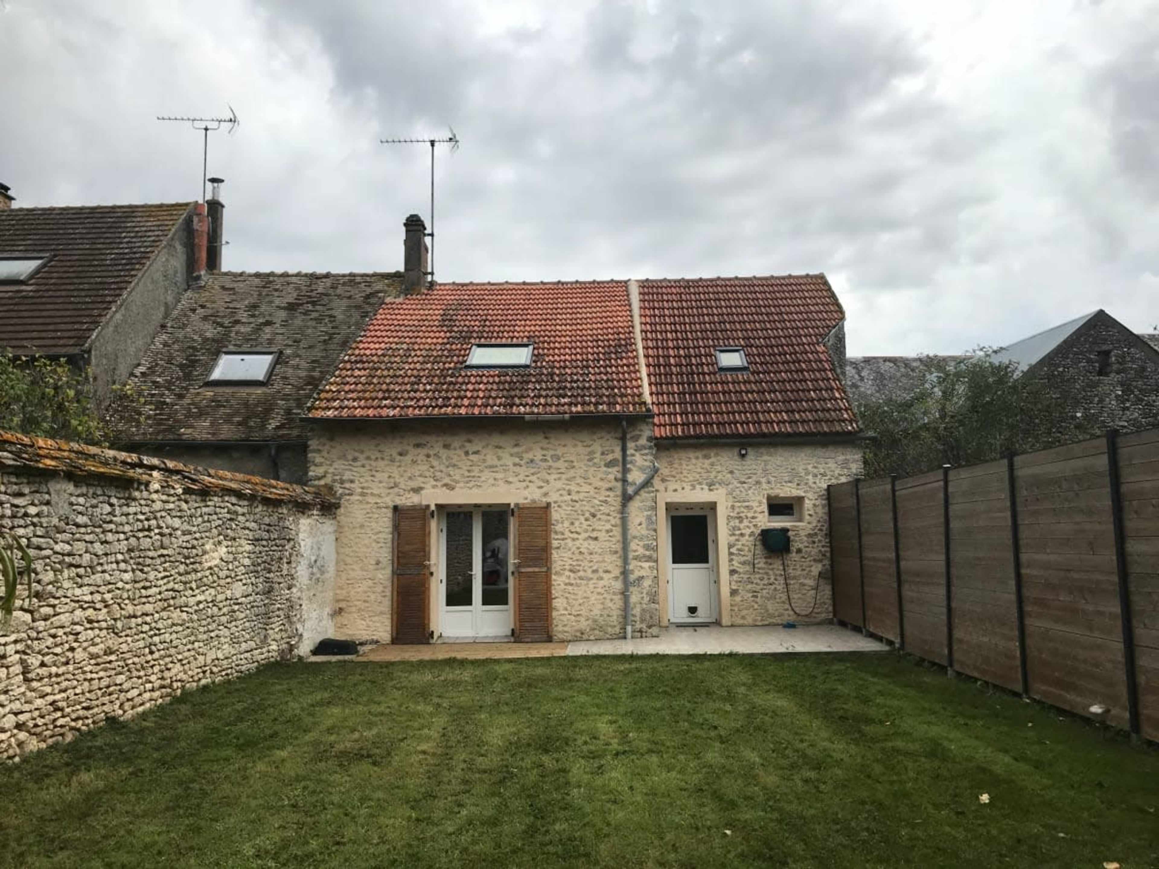 The image shows a stone house with a red-tiled roof, set in a yard surrounded by a wooden fence and neighboring homes.