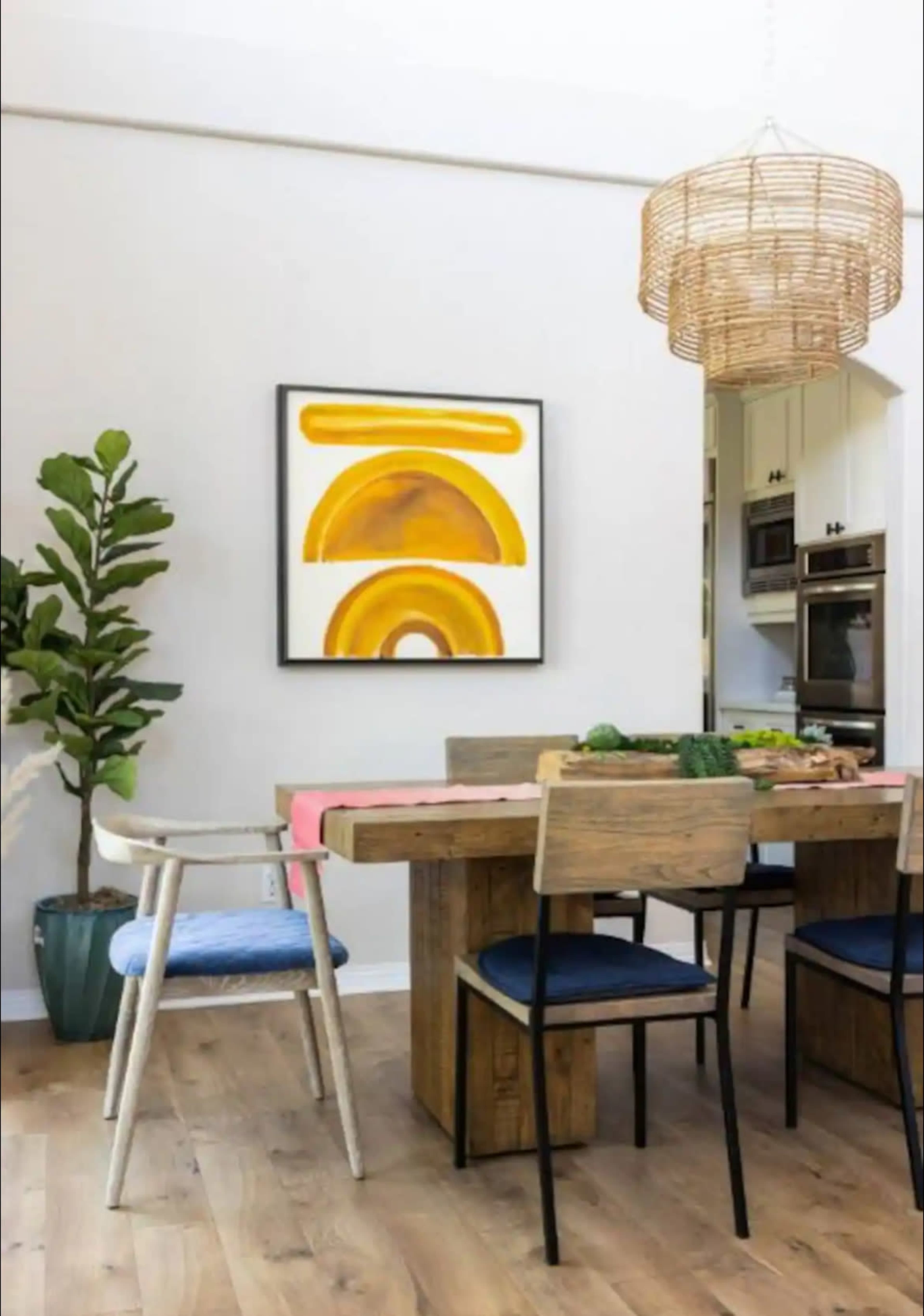 A wooden dining table with various vegetables sits in a bright room featuring a large abstract painting and a woven light fixture.