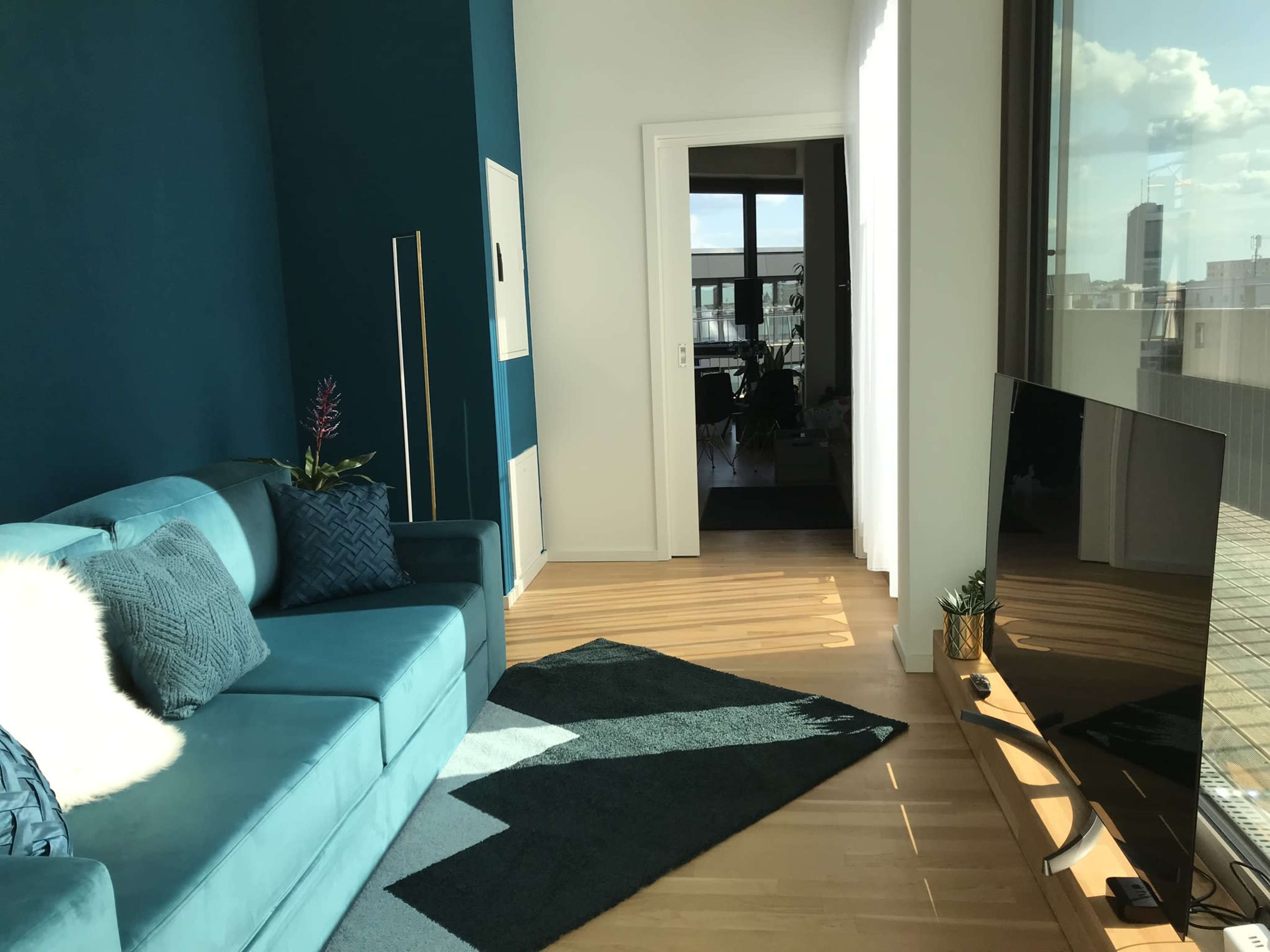 A modern living room with a turquoise sofa, a TV stand, and a partially visible hallway leading to another room.