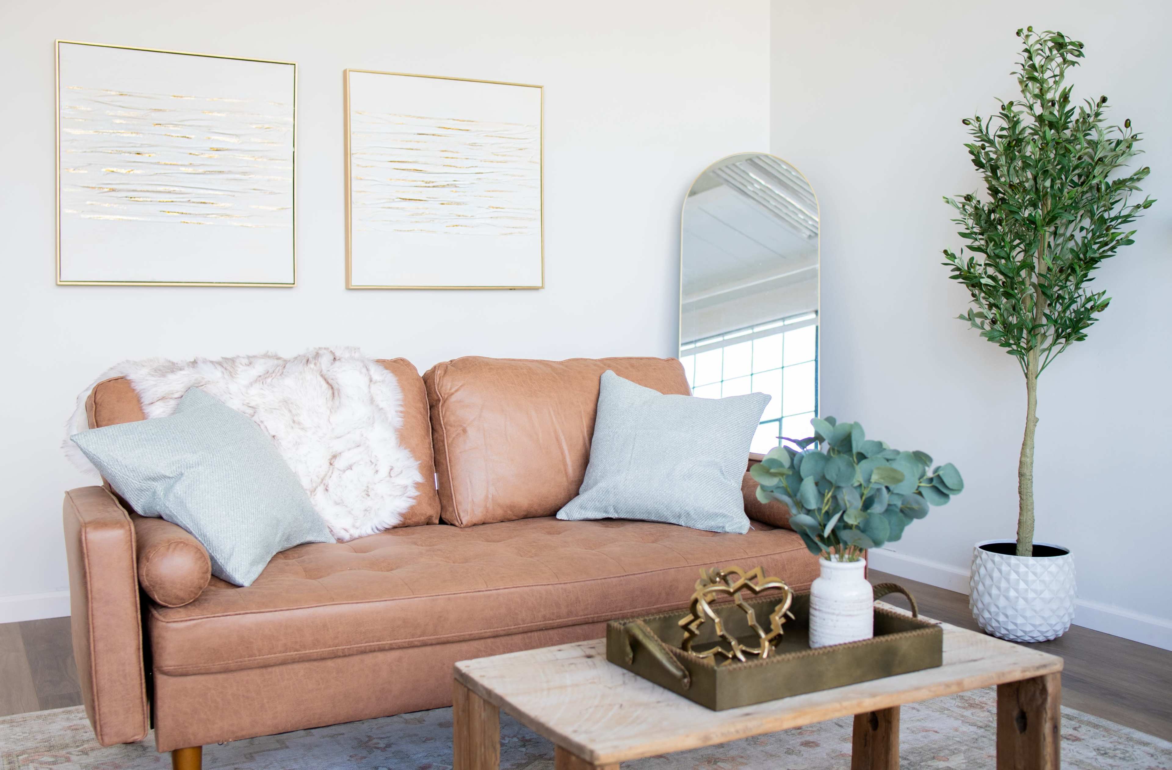 The image features a light brown couch adorned with blue pillows, accompanied by a wooden coffee table holding decorative items, a tall indoor plant, and two framed artworks above the couch.