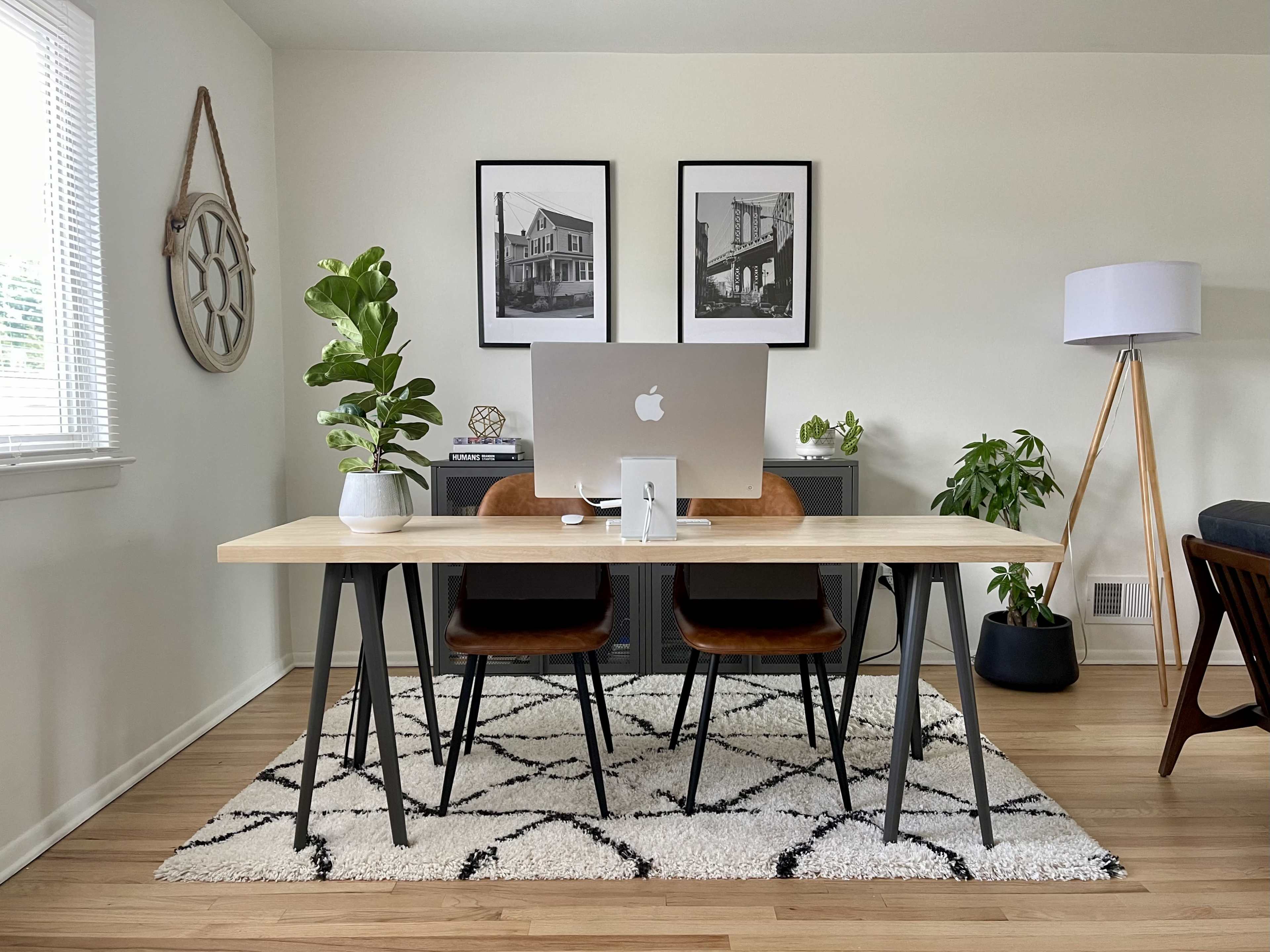 A modern home office setup featuring a wooden table with a desktop computer, two chairs, decorative plants, framed photographs on the wall, and a standing lamp.
