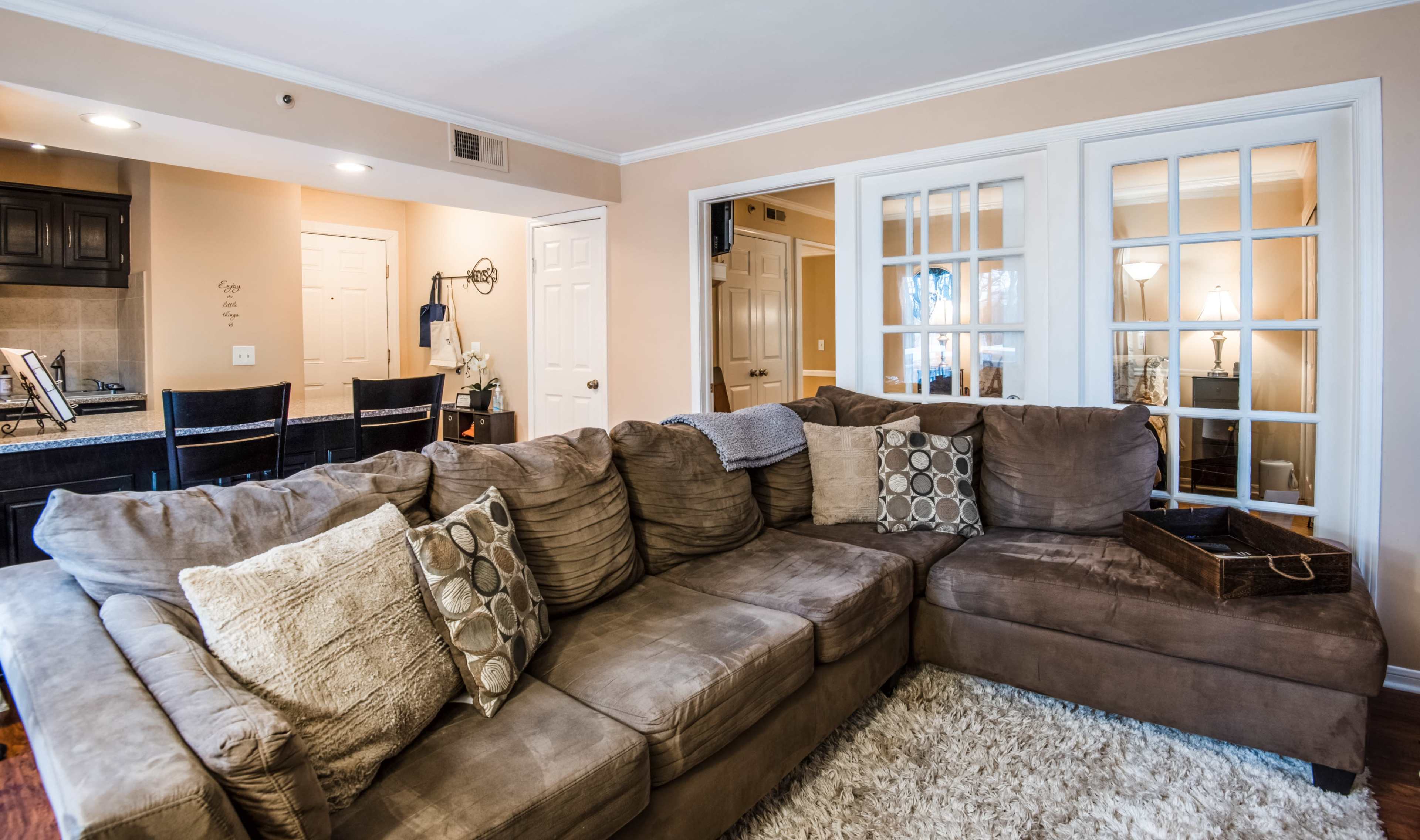 A spacious living area featuring a large, sectional brown couch with decorative pillows, next to a dining area and kitchen visible through a set of French doors.