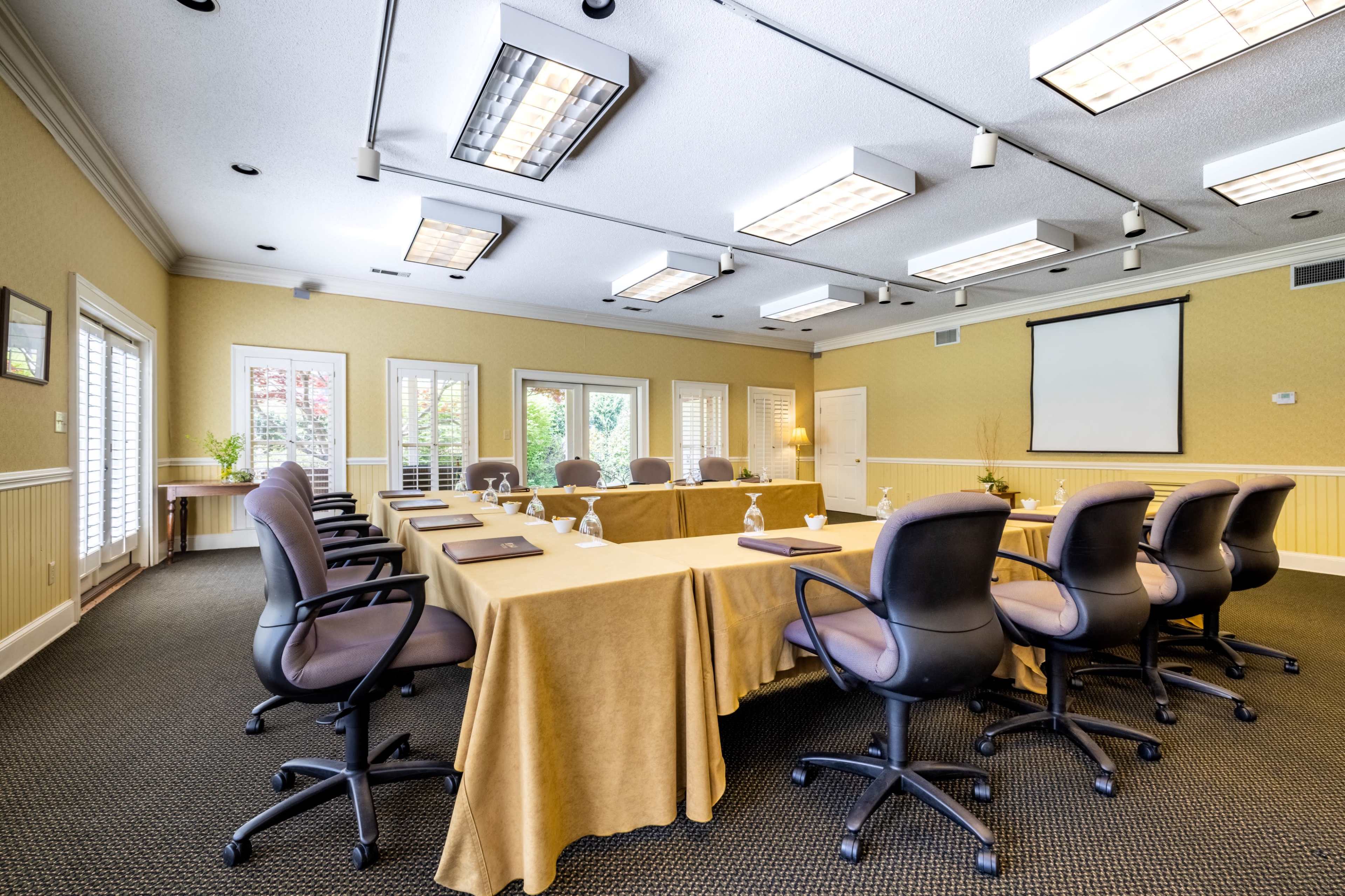 A conference room features a rectangular table surrounded by rolling chairs, with large windows allowing natural light and a screen at one end of the room.