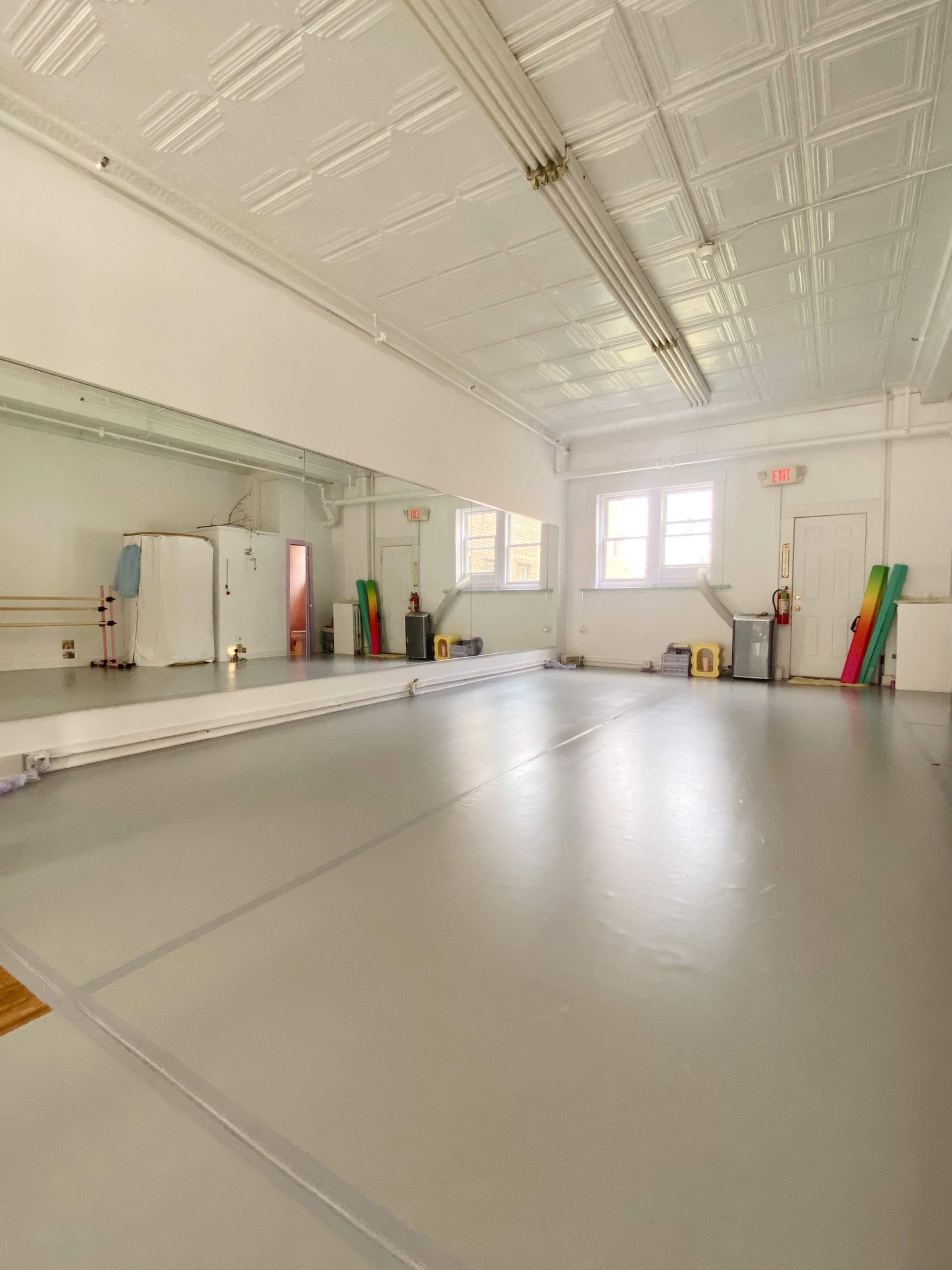 The image shows a spacious, empty dance studio with a mirrored wall and gray flooring.