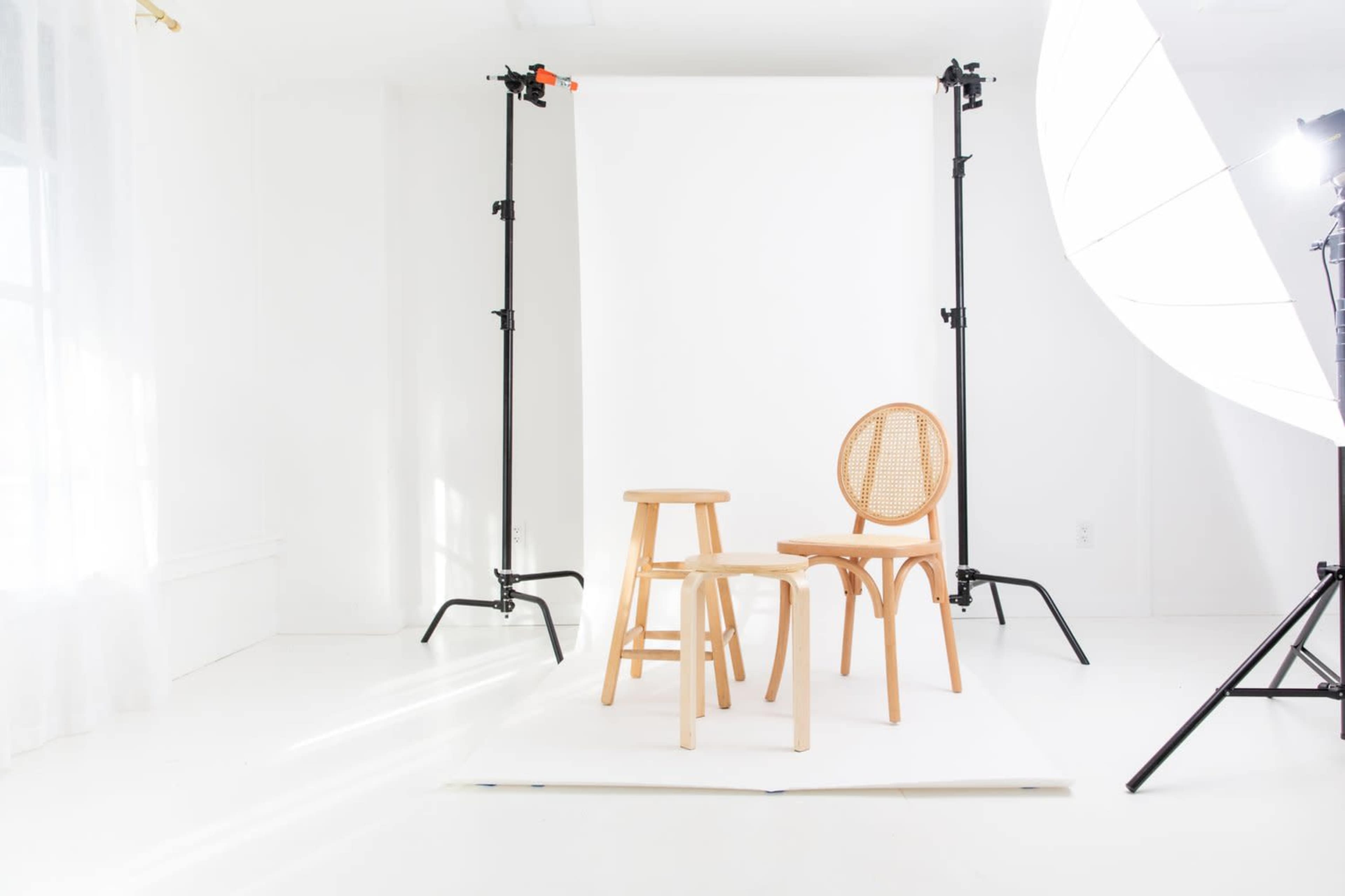 A bright, minimalist photography studio setup with two wooden chairs and a stool positioned in front of a white backdrop.