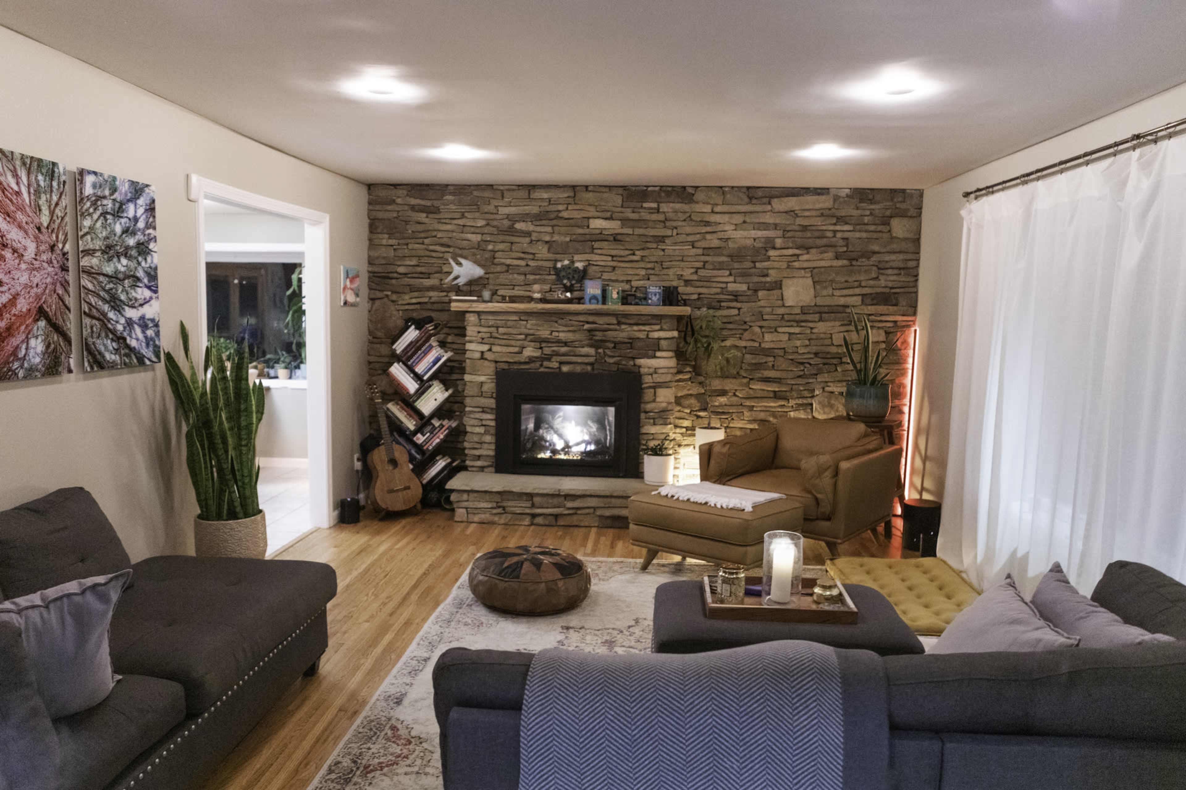 The living room features a stone fireplace surrounded by plants, comfortable seating, and a mix of decorative elements including artwork and a cozy area rug.