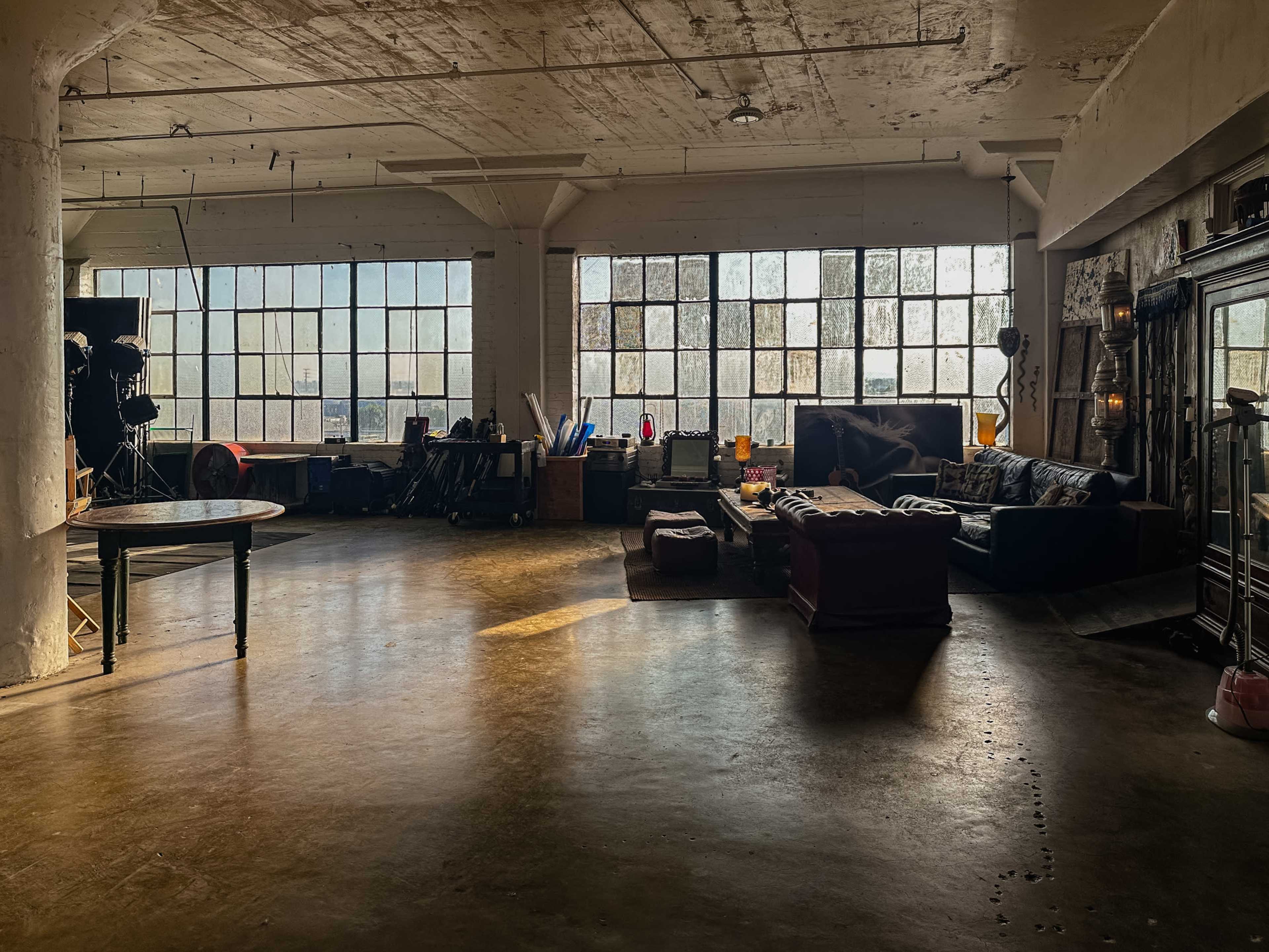 A spacious industrial loft features large windows, casting light onto various pieces of furniture and scattered props throughout the room.