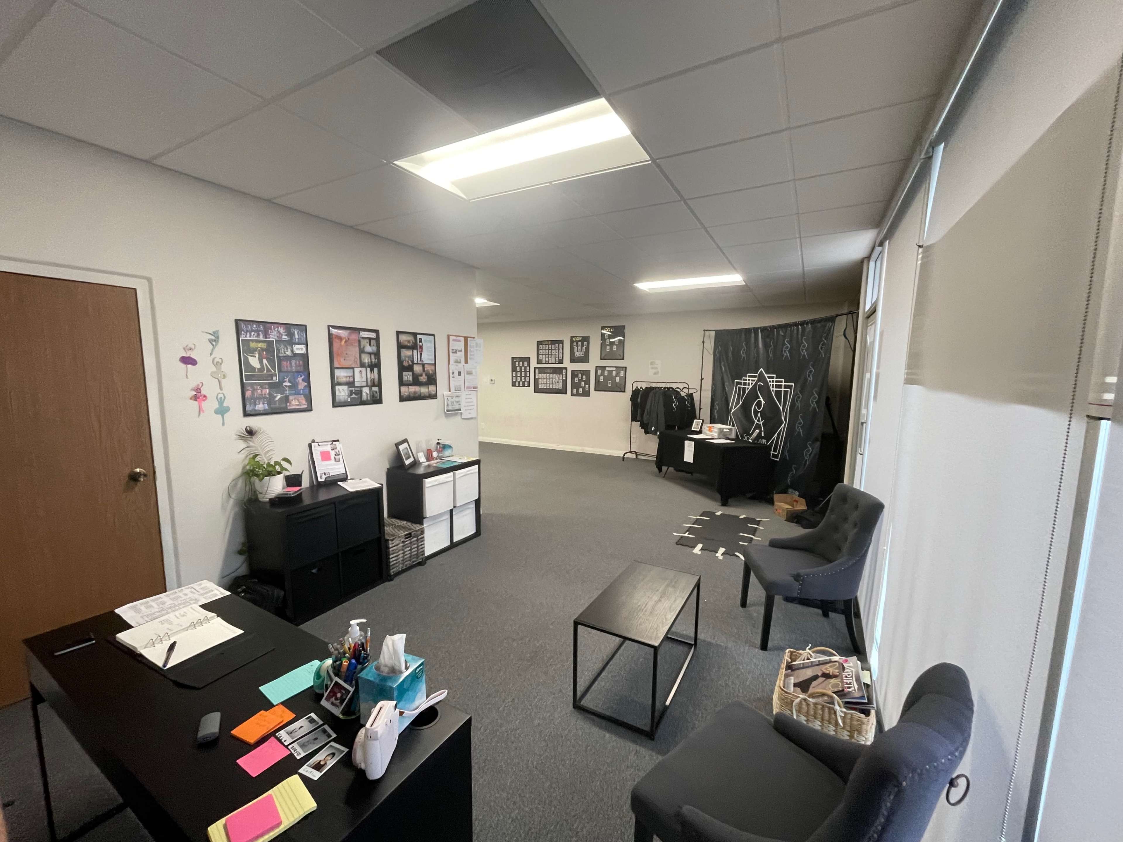 The image shows a spacious office area featuring black furniture, various framed photographs on the walls, and a clothing rack in the background.