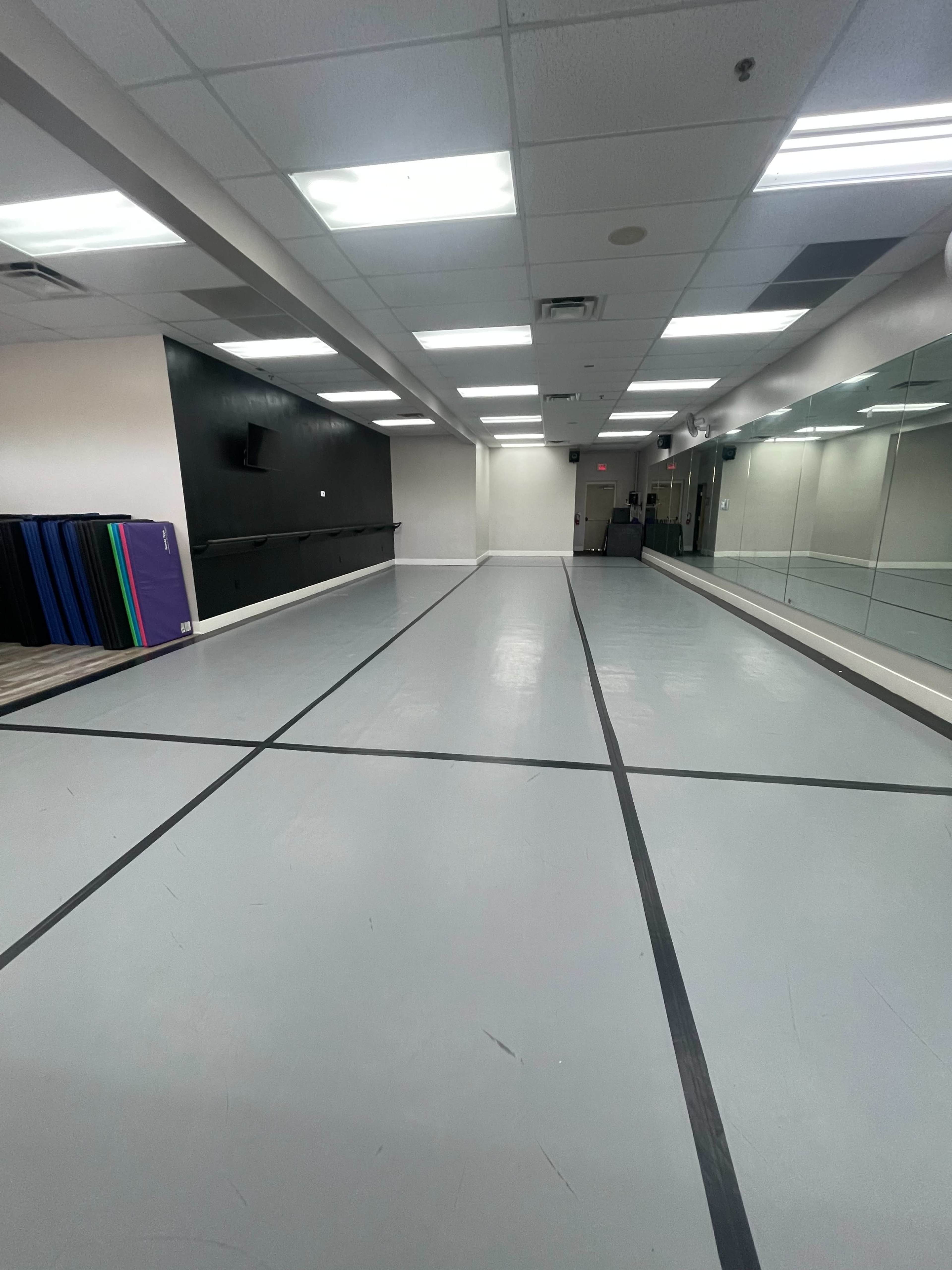 A spacious, empty dance studio with a mirrored wall and a grey floor marked by black lines.