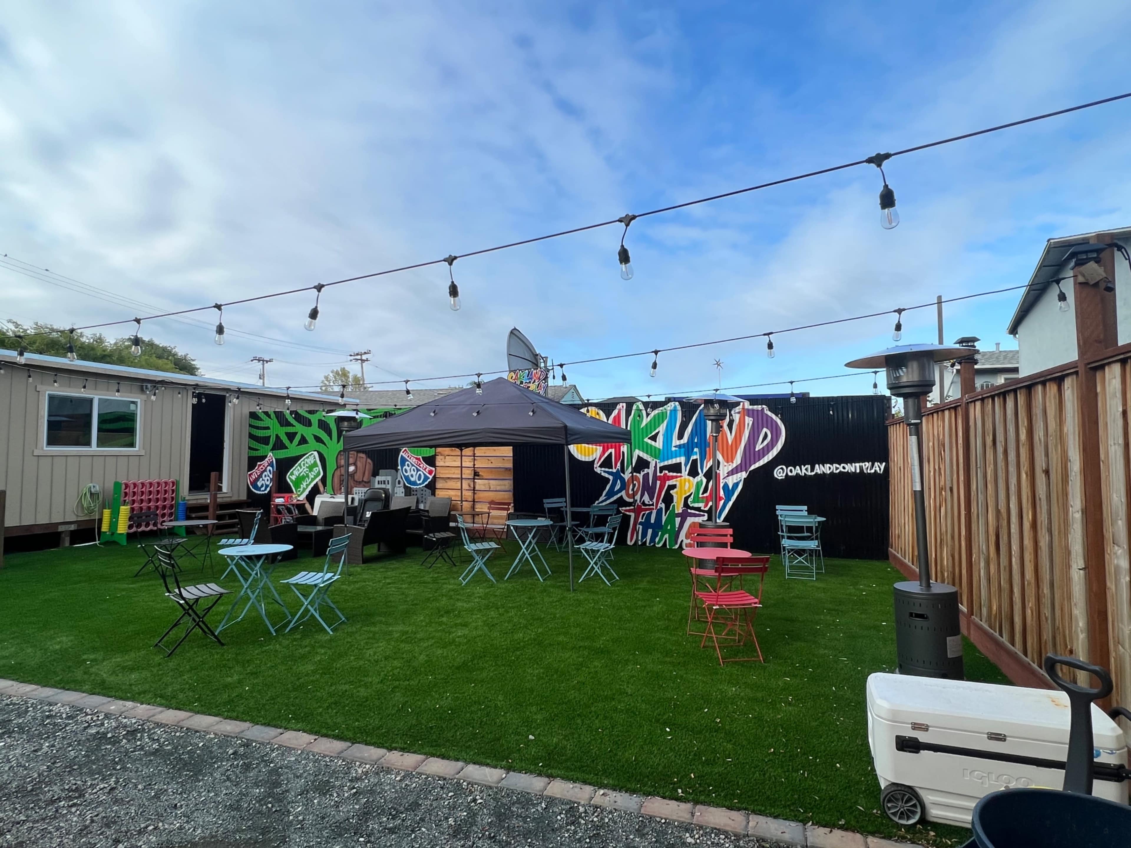 The scene features an outdoor social area with grassy turf, string lights, colorful chairs and tables, and graffiti on the walls in a setting conducive to gatherings.
