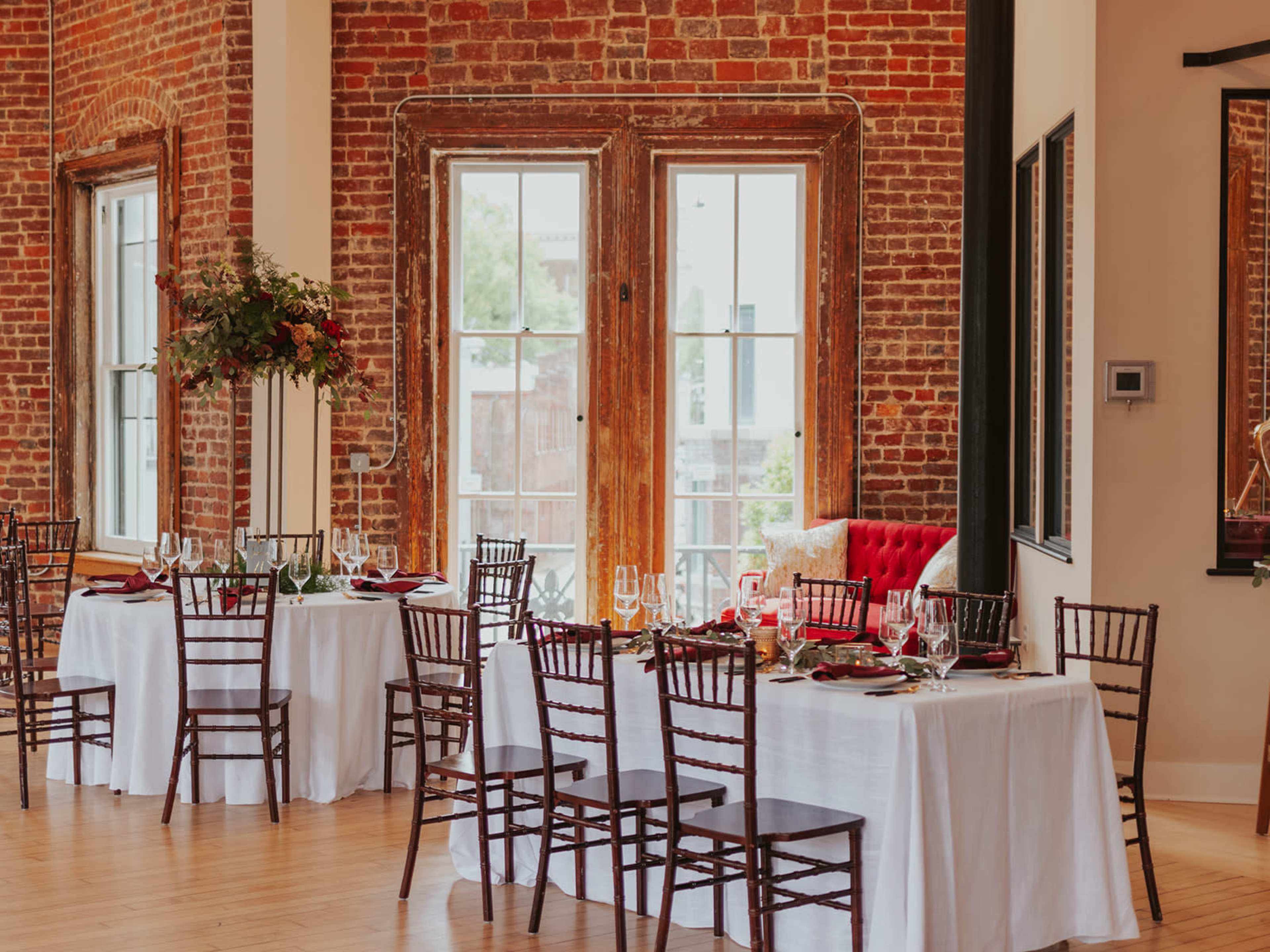 The interior features a dining area with round tables covered in white tablecloths, surrounded by wooden chairs, and exposed brick walls, illuminated by natural light from large windows.