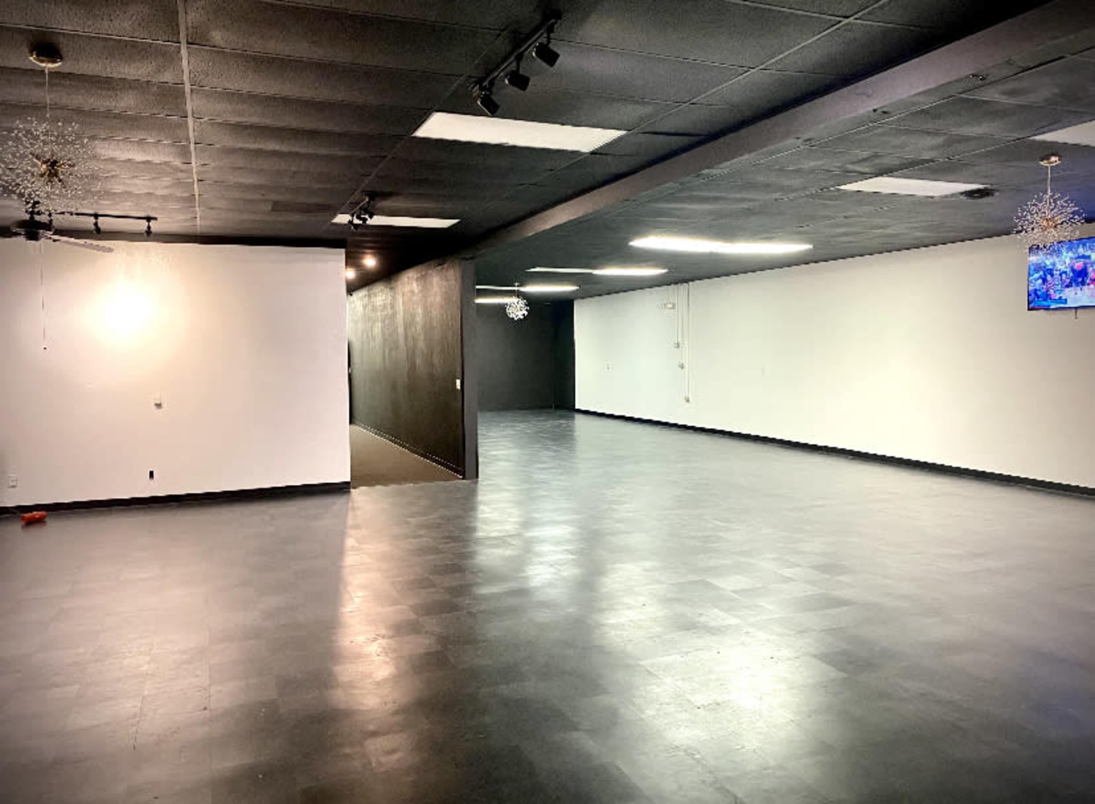 The image shows a large, empty room with black flooring and a dark wall, illuminated by overhead lights.