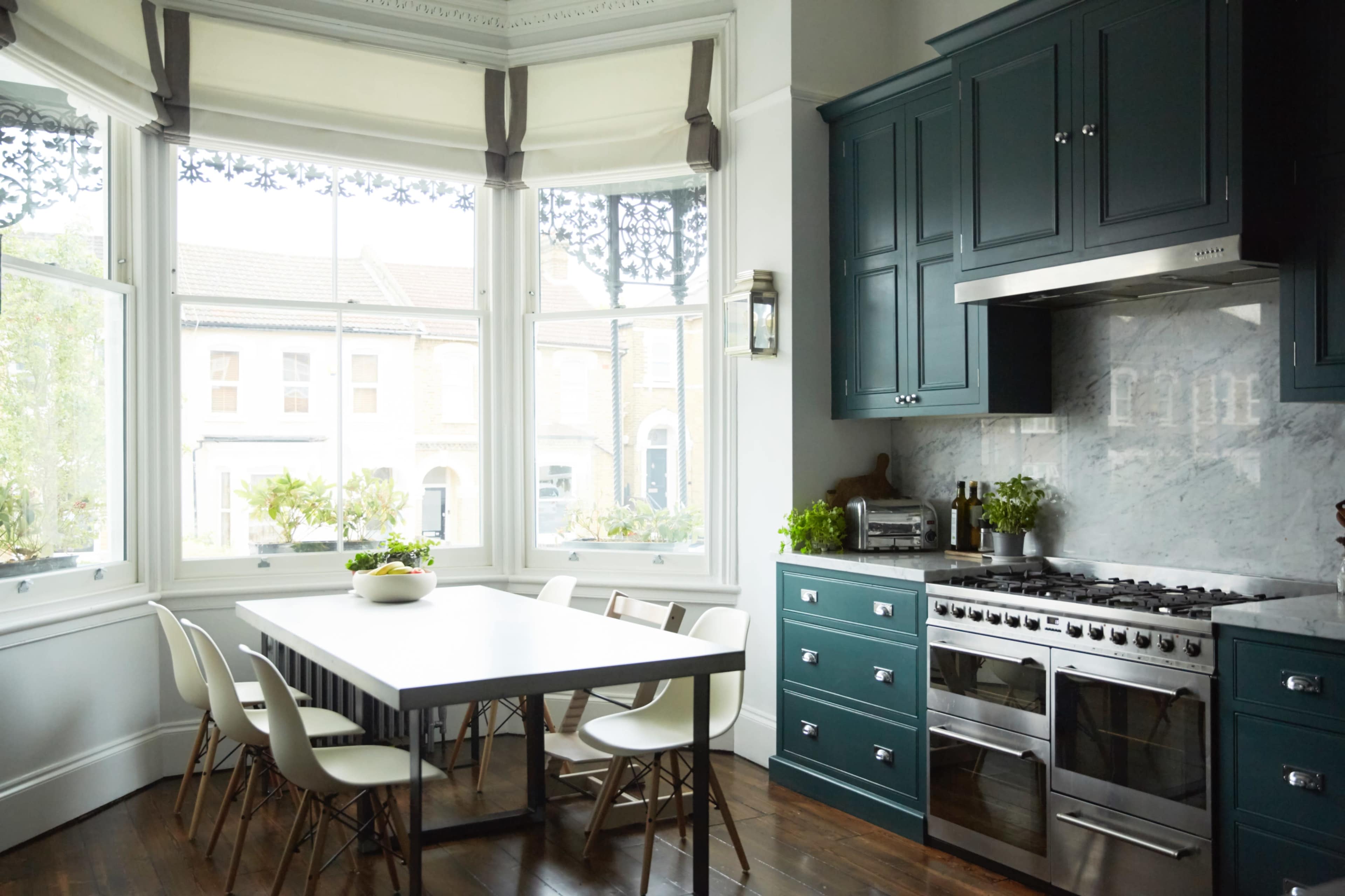 A modern kitchen features dark green cabinetry, a stainless steel stove, and a dining table surrounded by white chairs, with large windows allowing natural light in.