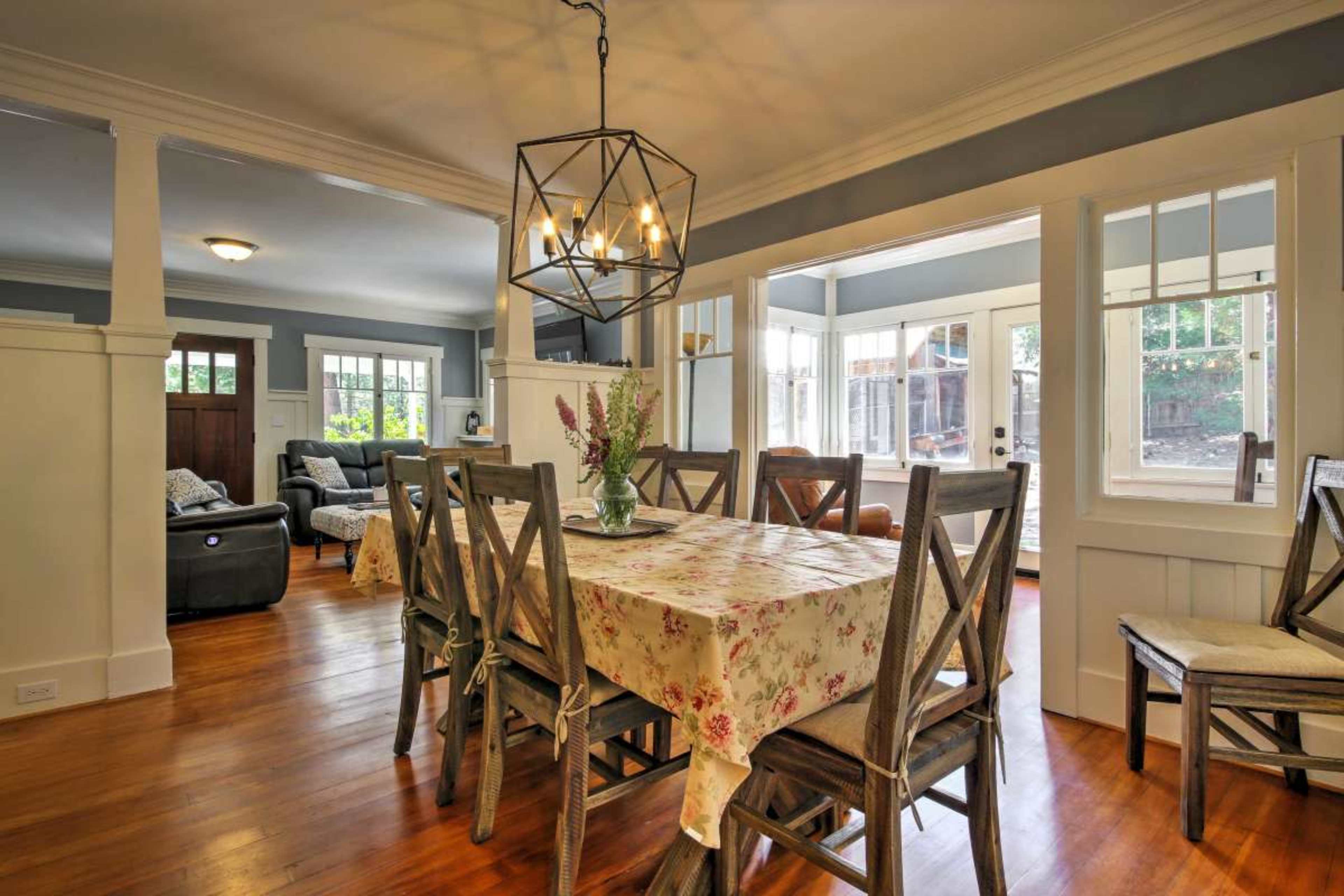 A dining area features a wooden table surrounded by chairs, with a geometric light fixture above and large windows showcasing a view of the outdoor space.