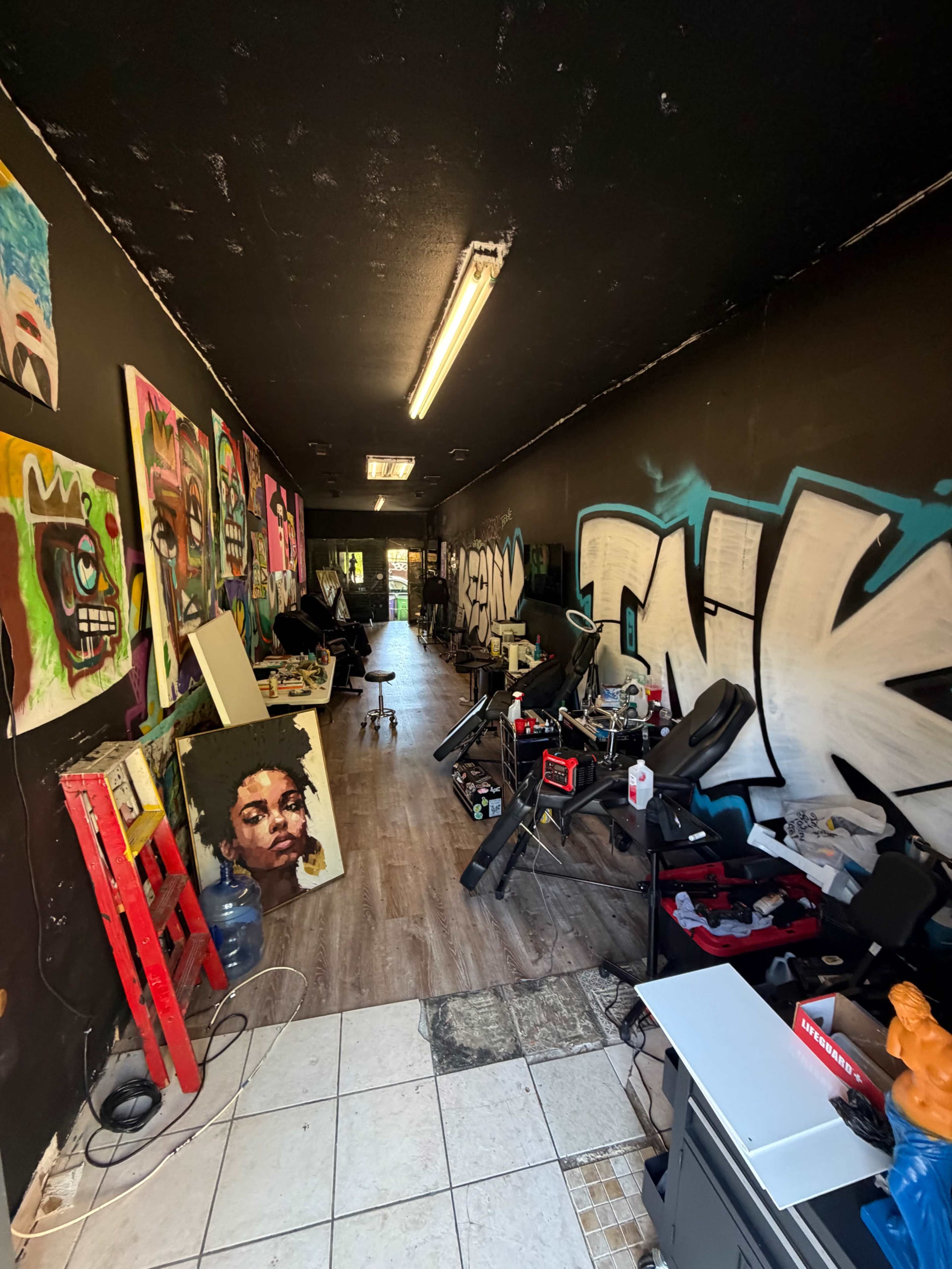 The image shows a narrow, dimly lit hallway in an art studio, lined with vibrant graffiti and artworks on the walls, with various furniture and art supplies scattered throughout.