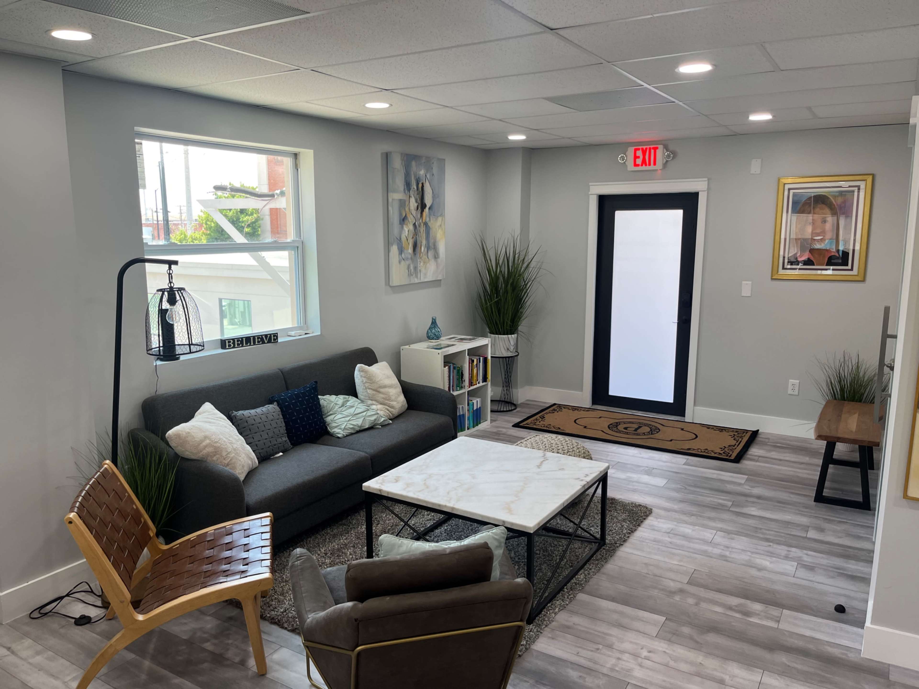 A modern, well-lit living area with a sofa, a coffee table, a couple of chairs, and plants, alongside a door leading outside.