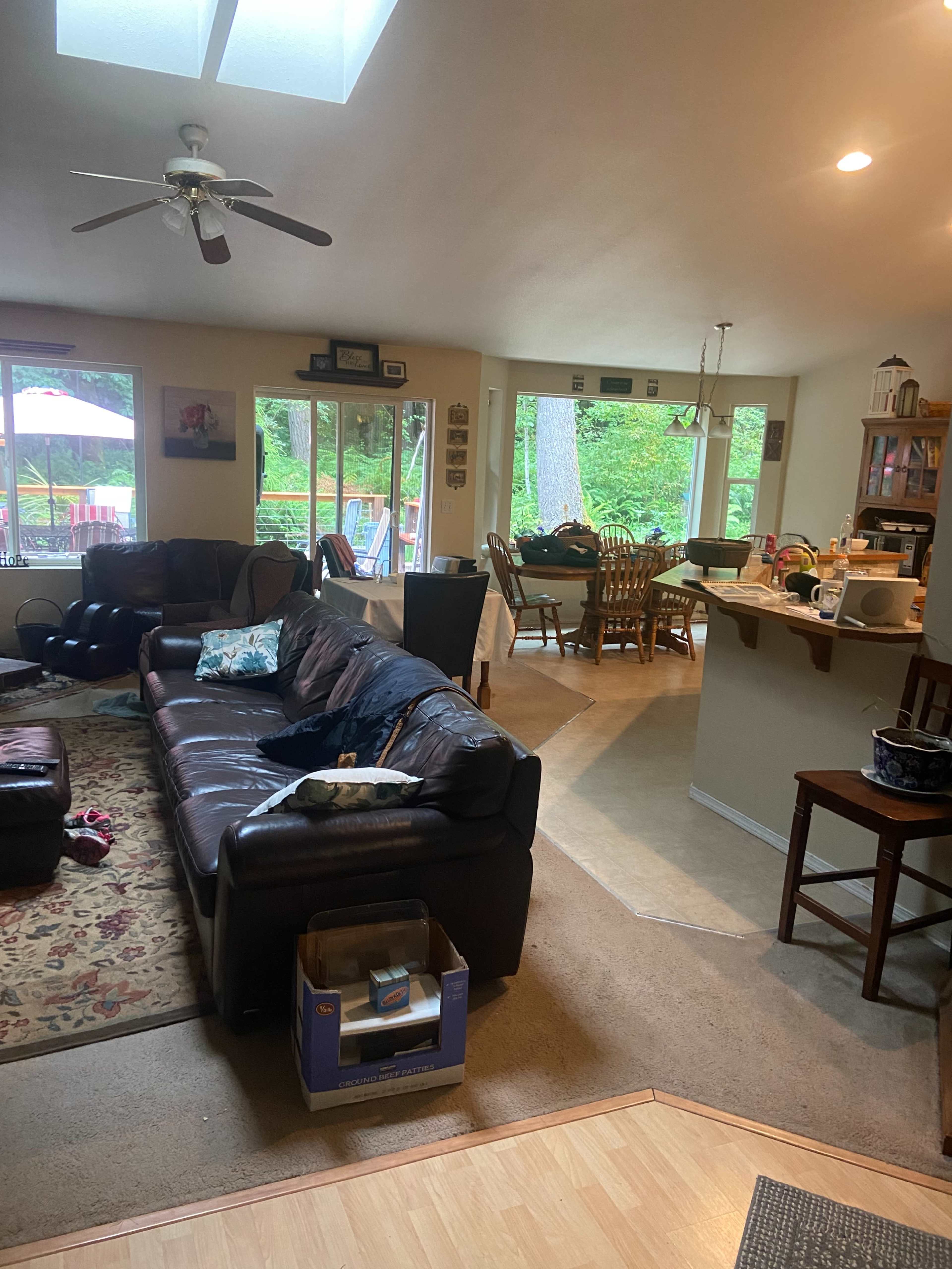 The interior of a home features a living room with a leather couch and a dining area visible in the background, surrounded by large windows looking out to a green yard.