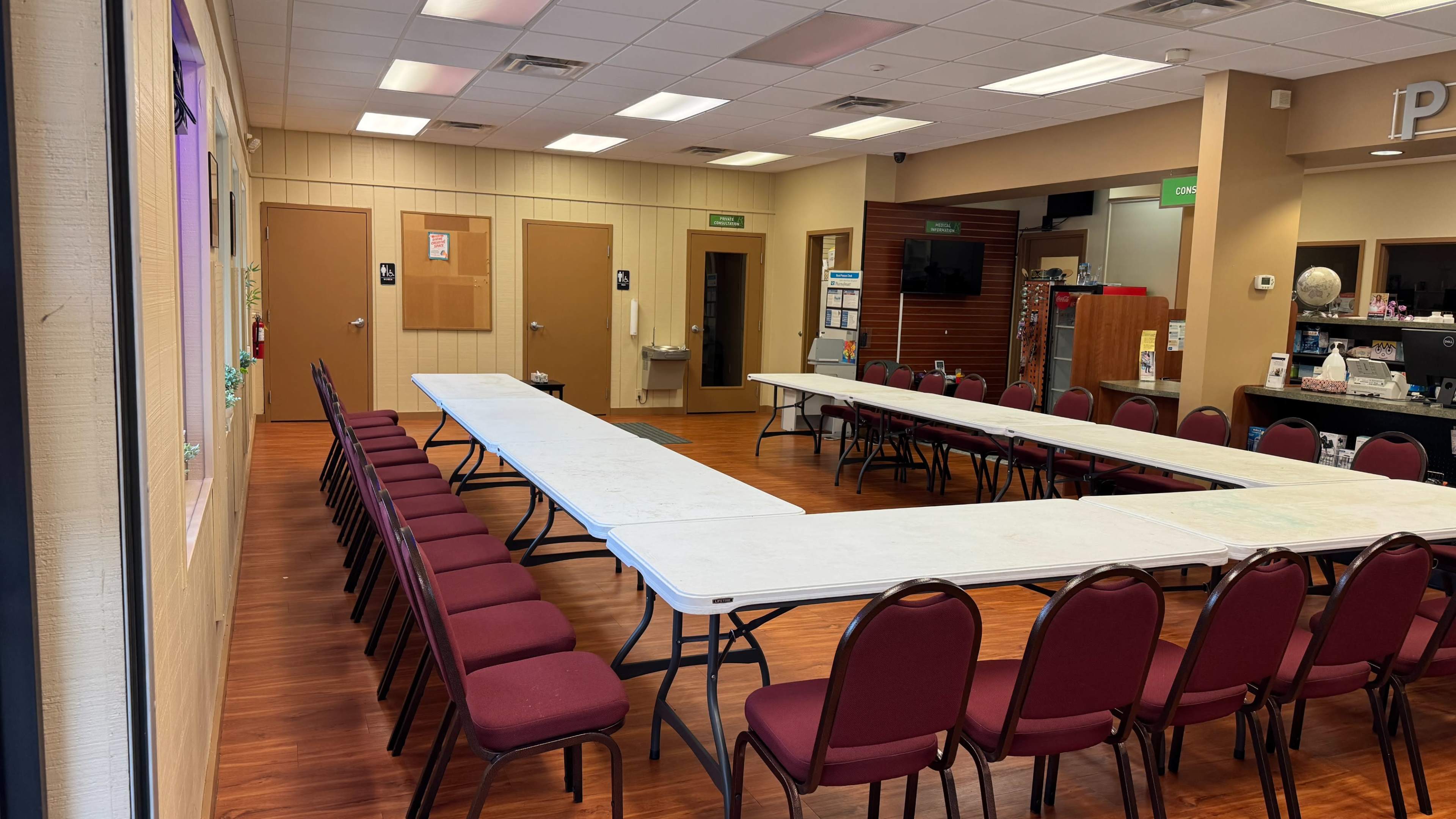 The image shows a spacious room arranged with several tables in a U-shape, surrounded by chairs, with walls featuring doors and informational displays.