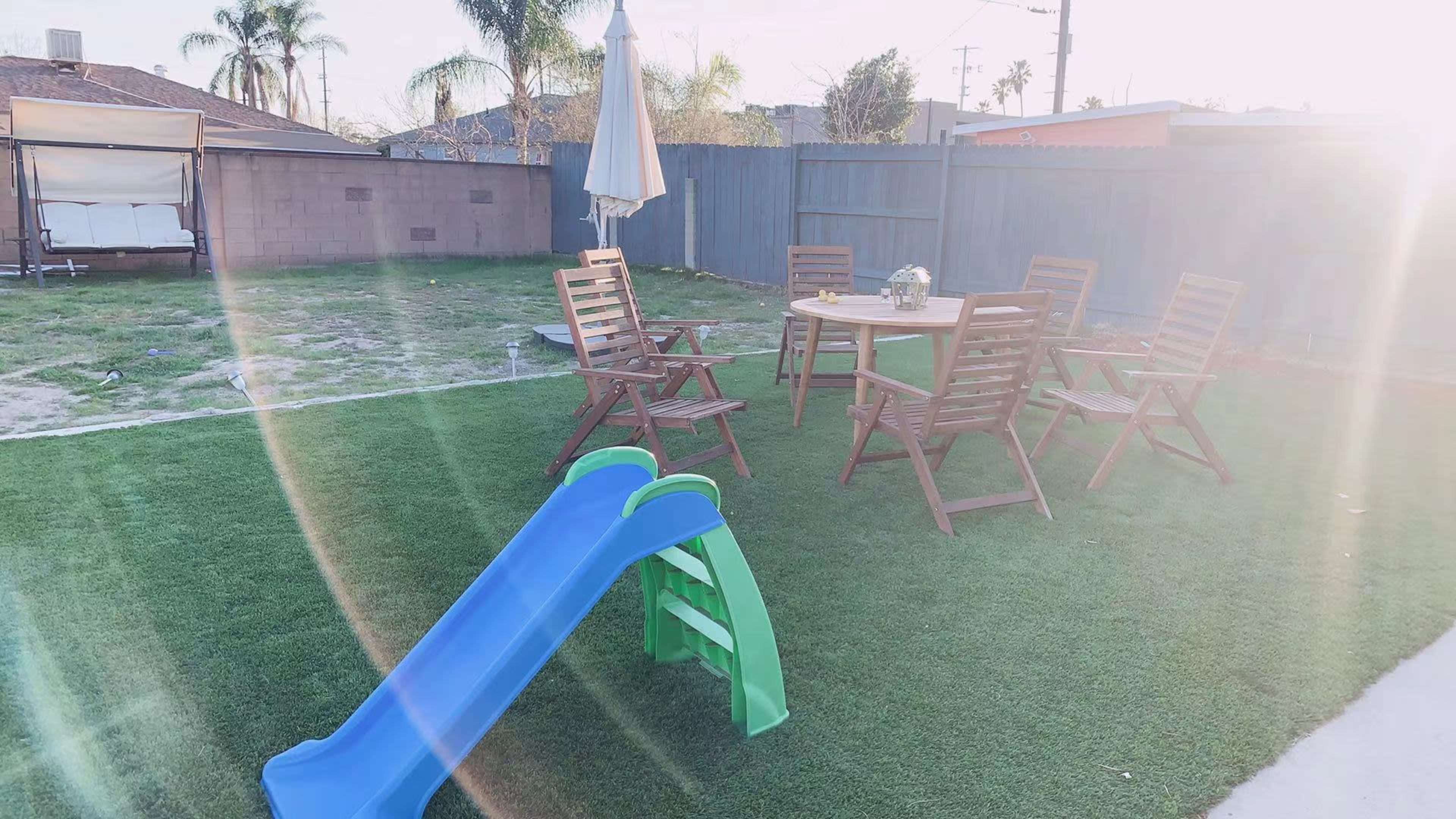 A small patio area features a table and four wooden chairs, with a blue and green slide on synthetic grass and a large umbrella nearby.