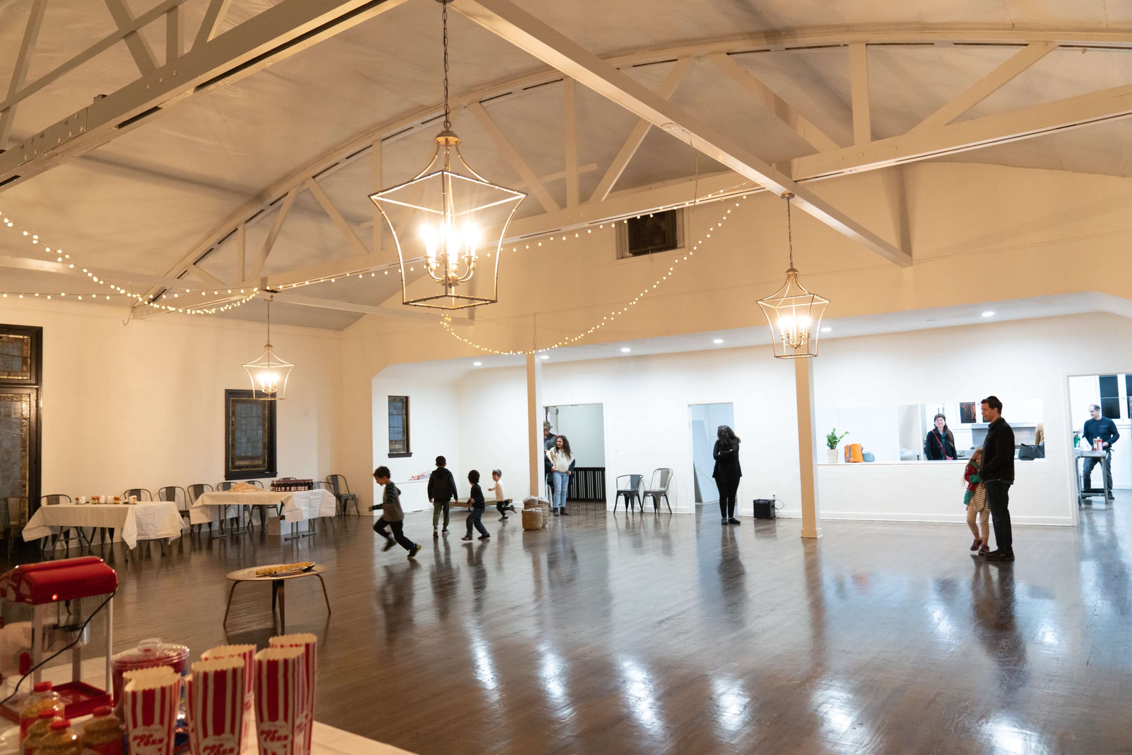 An indoor event space features a high ceiling with pendant lights, tables set along the perimeter, and a few guests in the background.