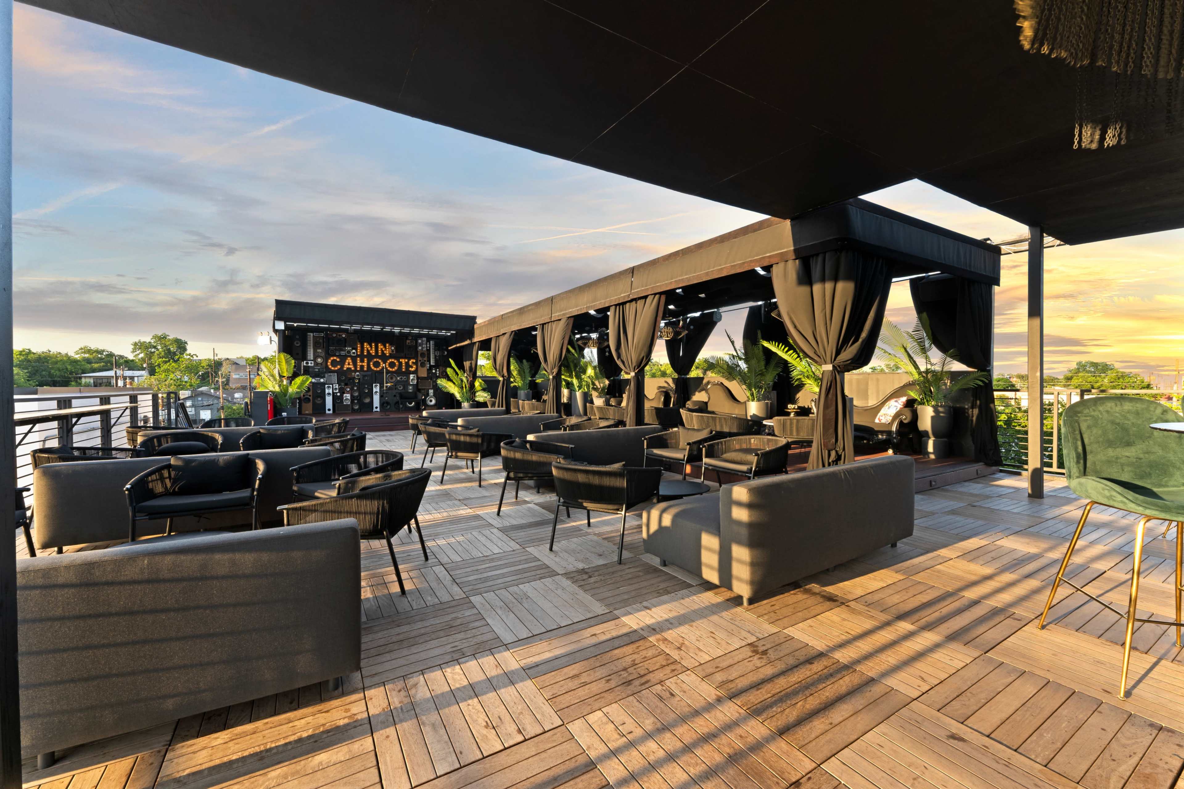 The image shows a rooftop patio with dark furniture, enclosed by curtains, and a bar named "Cahoots" in the background, set against a sunset sky.