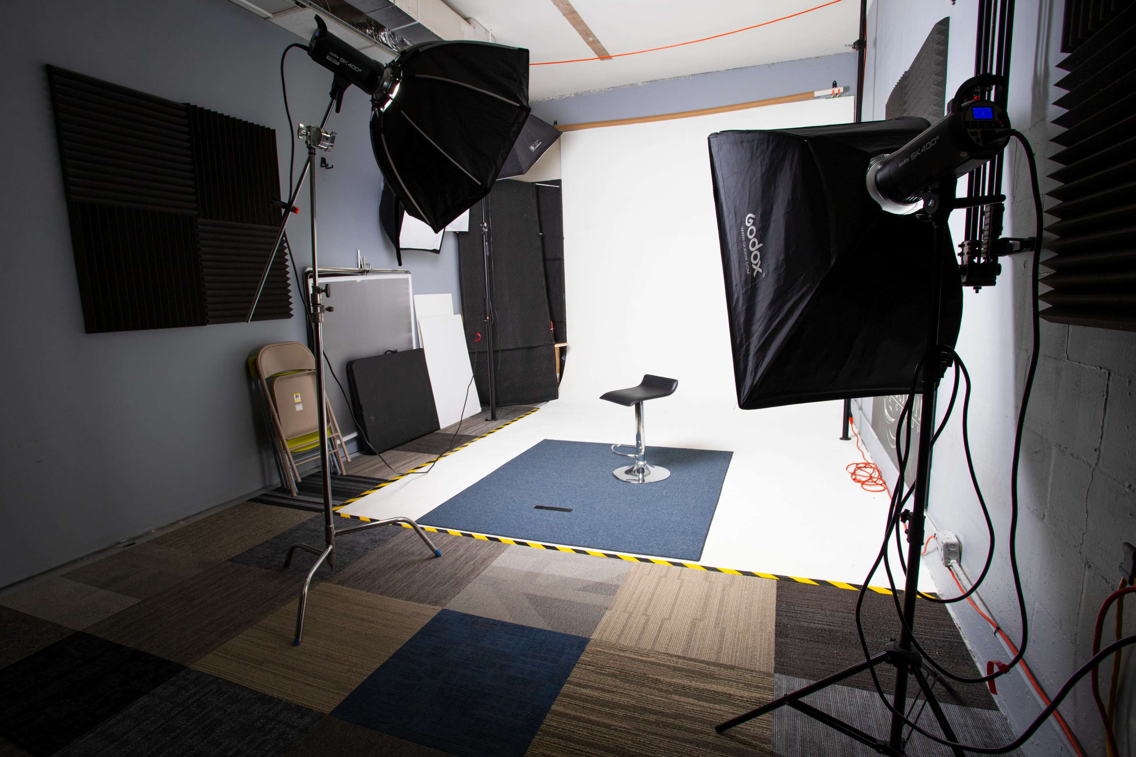 A photography studio is set up with two light sources, a black stool on a blue mat, and a white backdrop.