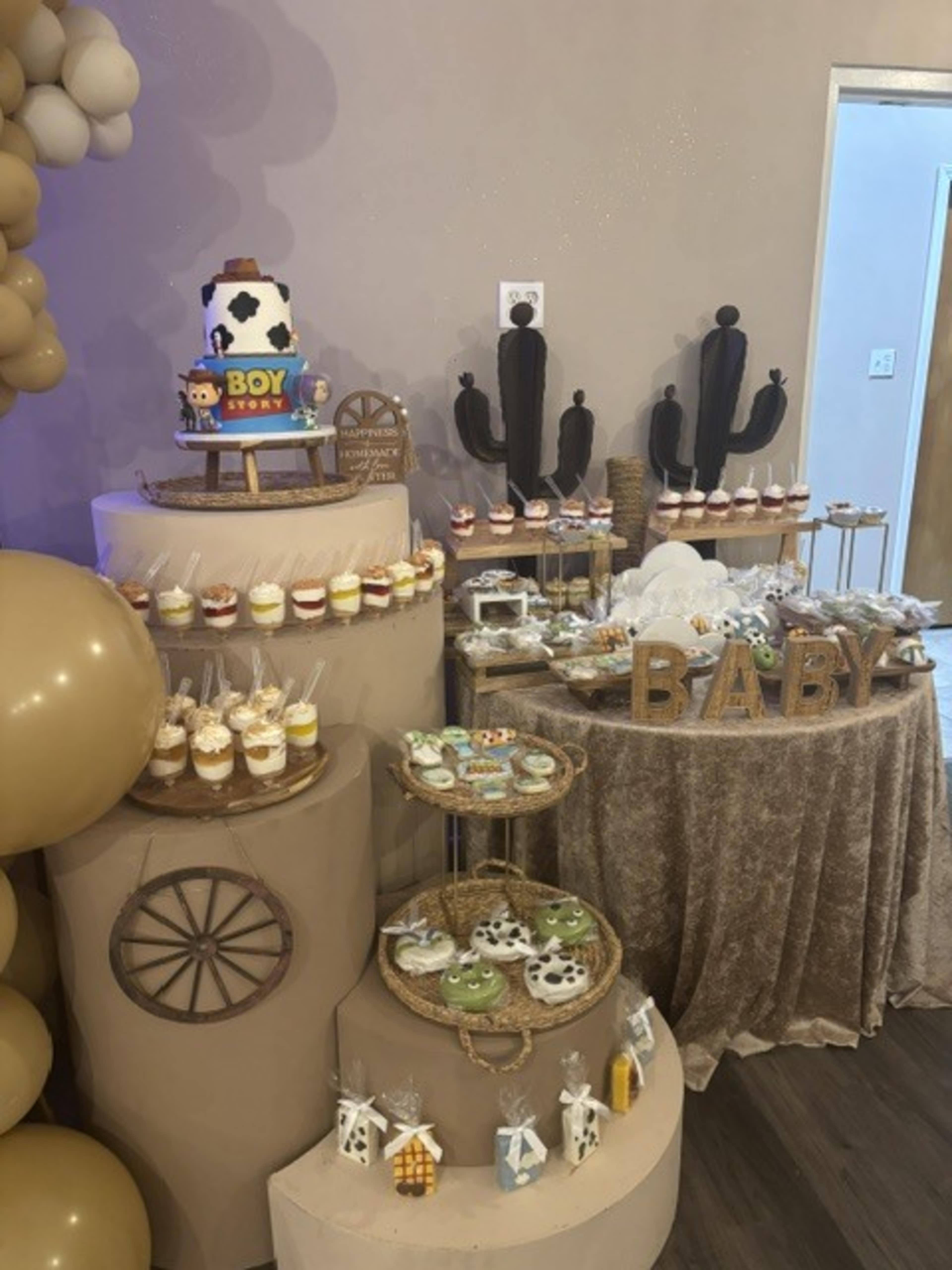 The image shows a dessert table decorated for a baby shower, featuring a tiered cake, various cupcakes, and themed treats arranged on multiple platforms.