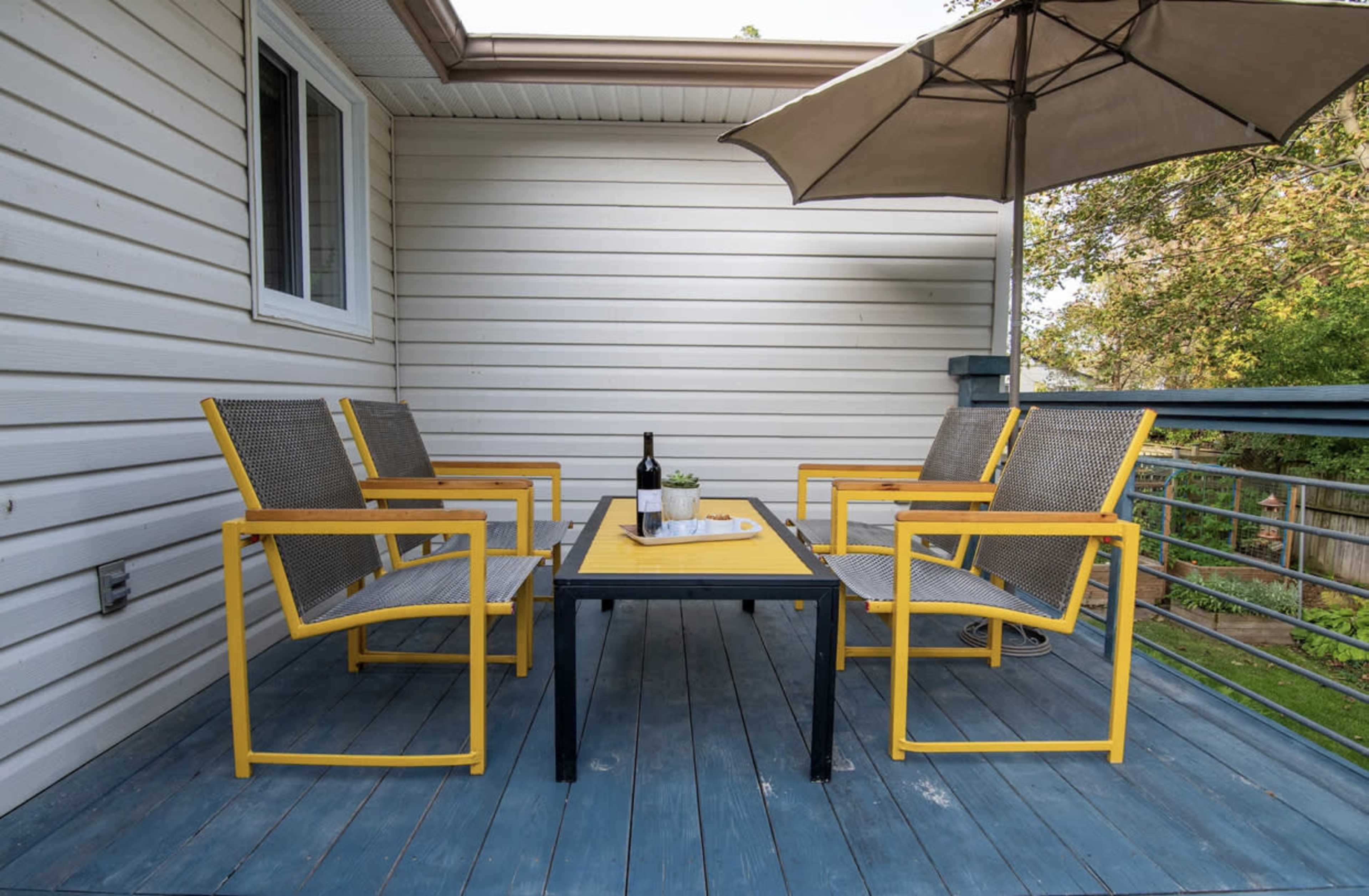 A small deck features four yellow and grey chairs surrounding a rectangular table with a bottle of wine and a plant, topped by a large umbrella.