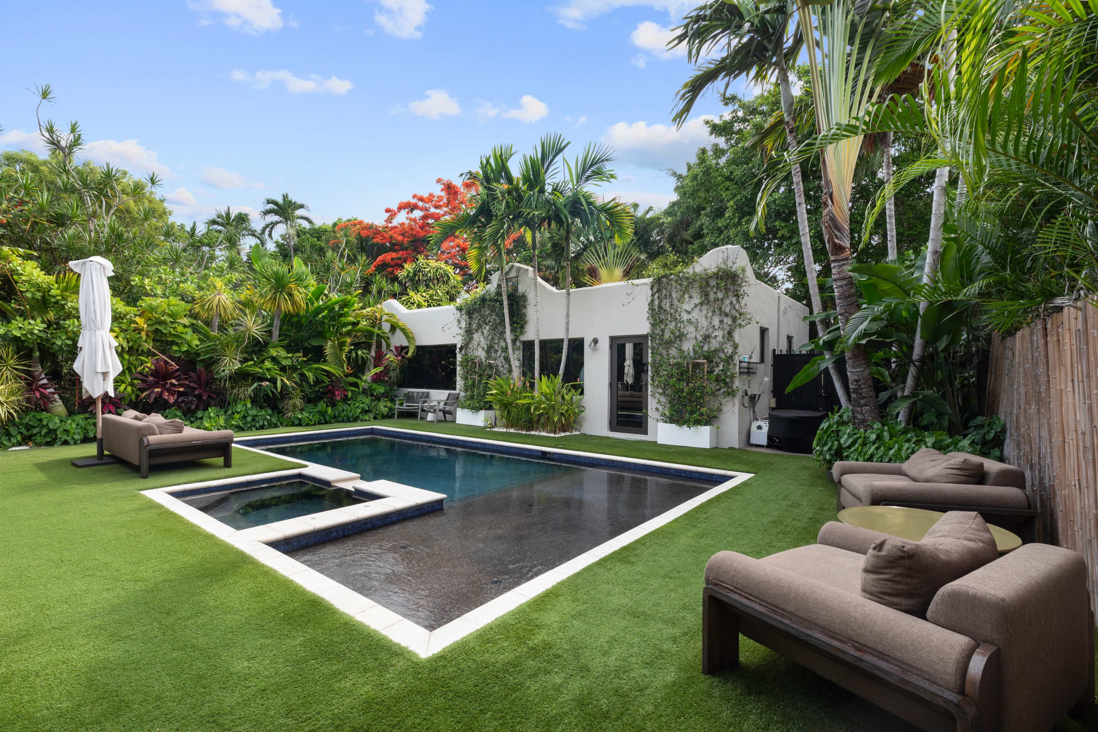 The image shows a modern backyard with a rectangular swimming pool surrounded by lush greenery and palm trees, along with two brown lounge chairs positioned on a grassy area.