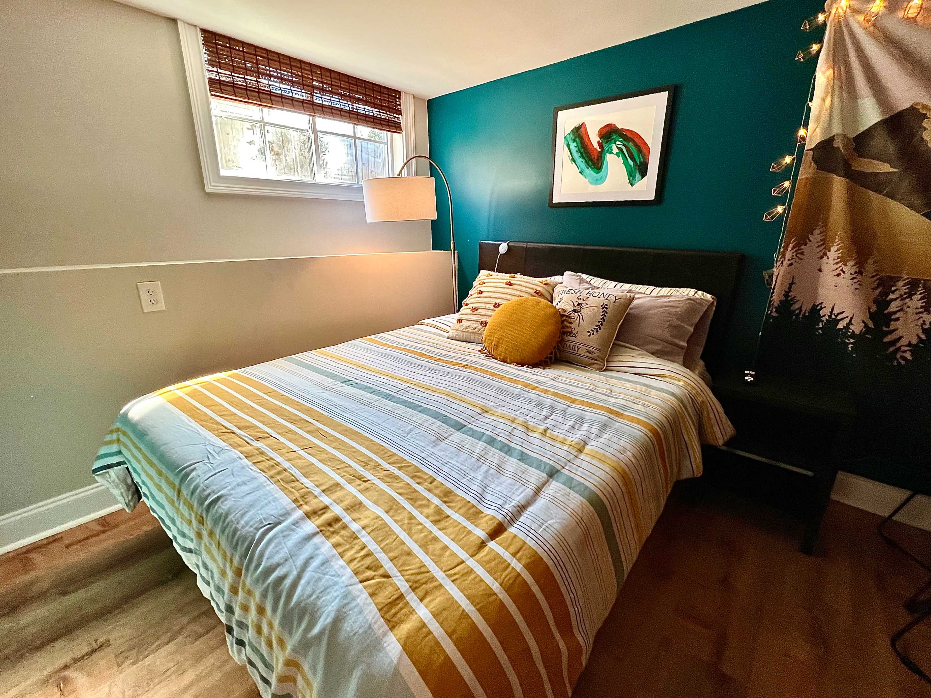 The image shows a well-lit bedroom with a double bed, featuring a striped comforter, decorative pillows, and a wall-mounted lamp beside a window.