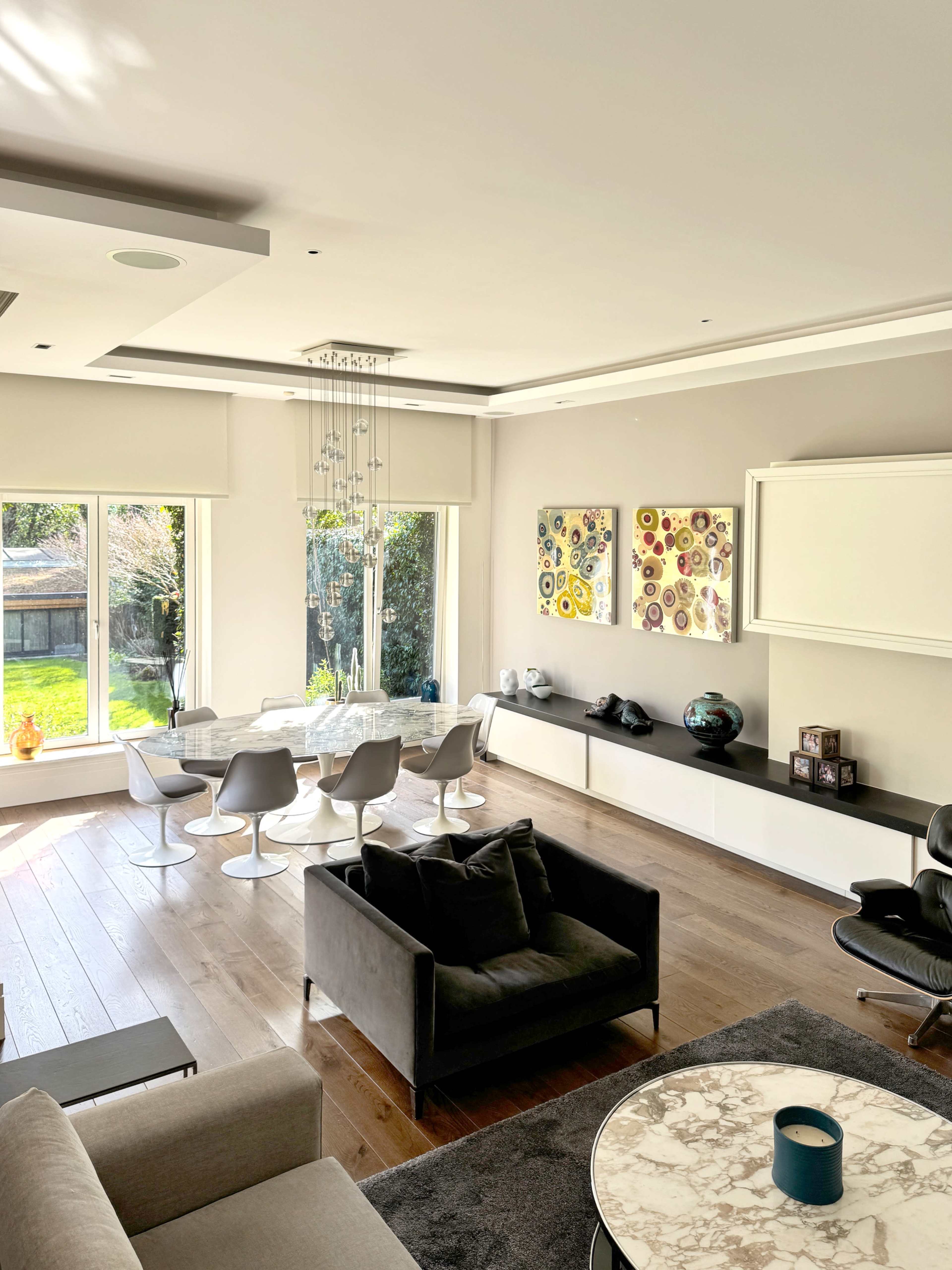 The image shows a bright living room featuring a dark sofa, a round marble coffee table, and modern chairs arranged around a glass table, with large windows overlooking a garden.