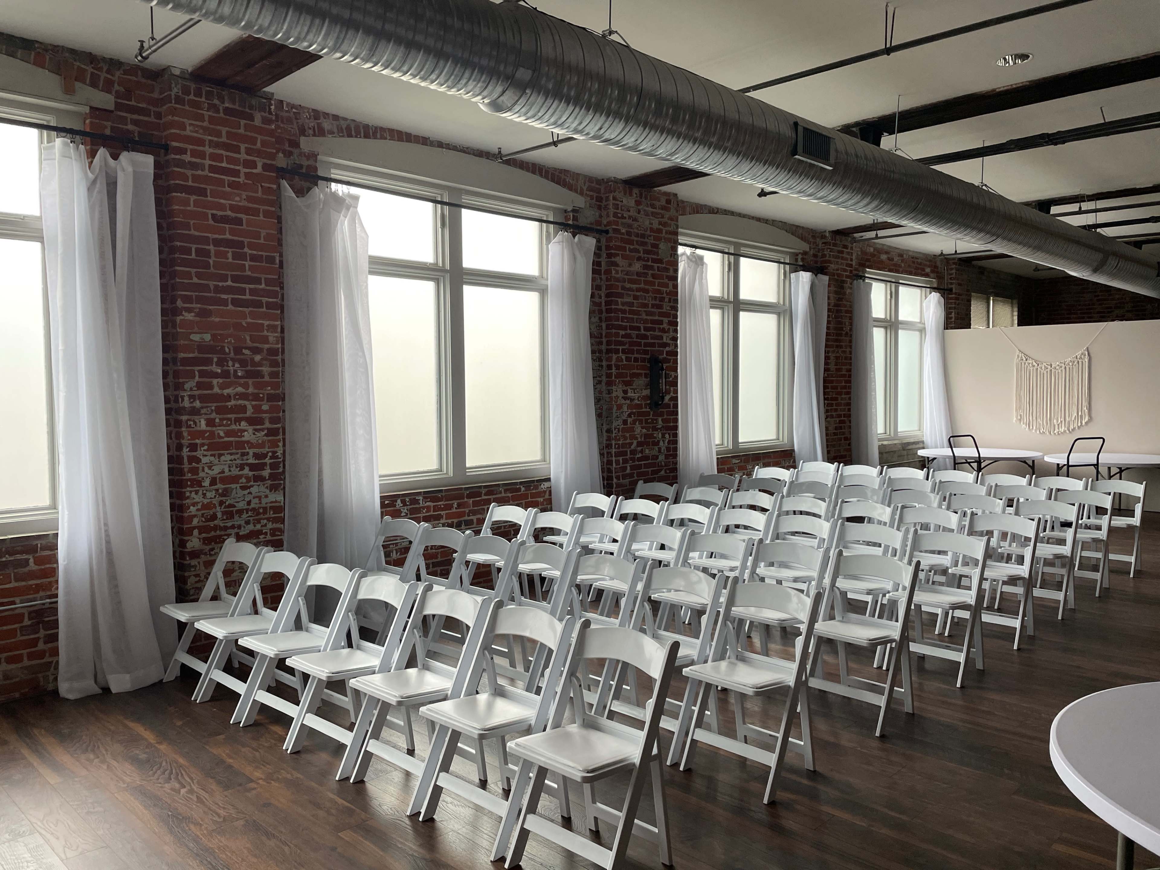 A row of white folding chairs is arranged in front of large windows in a room with exposed brick walls.