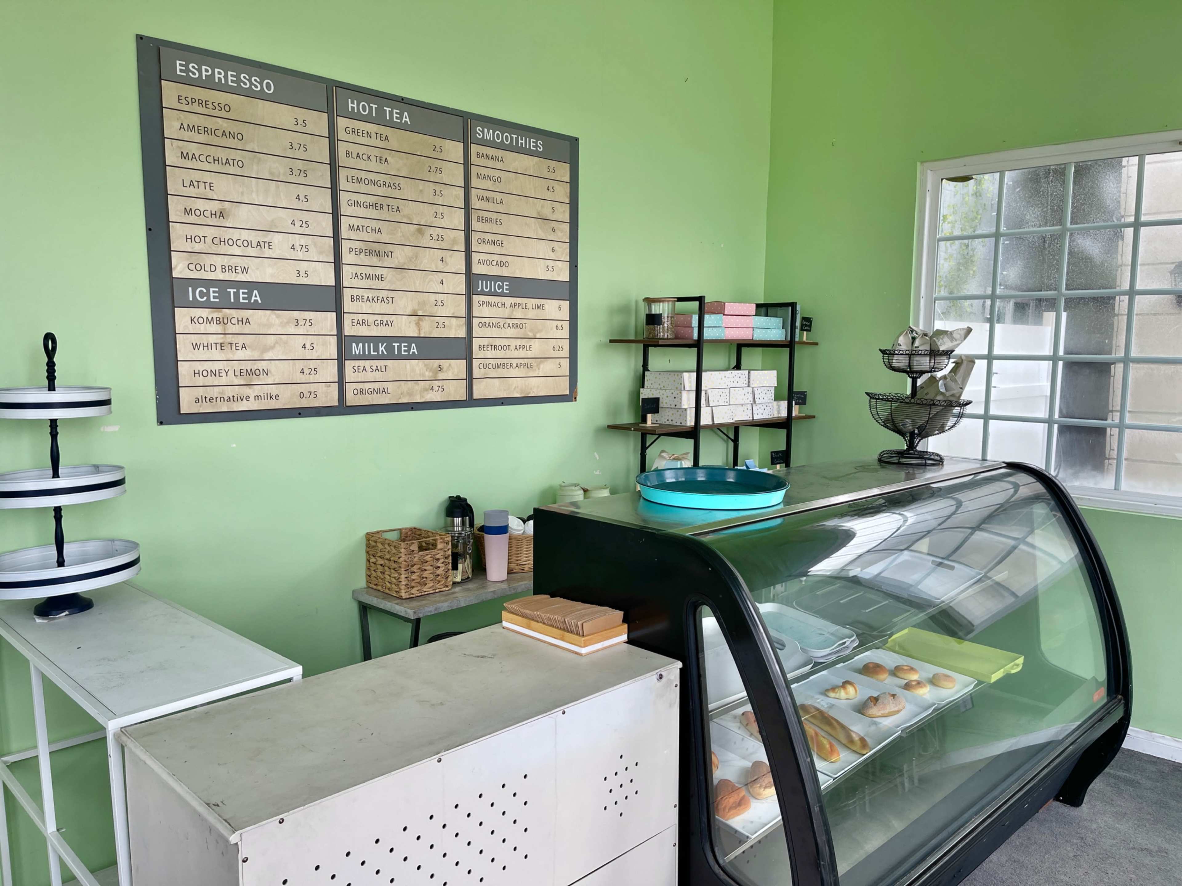 A cafe interior featuring a display case with pastries, a menu on the wall, and a green-painted wall with various shelves and containers.