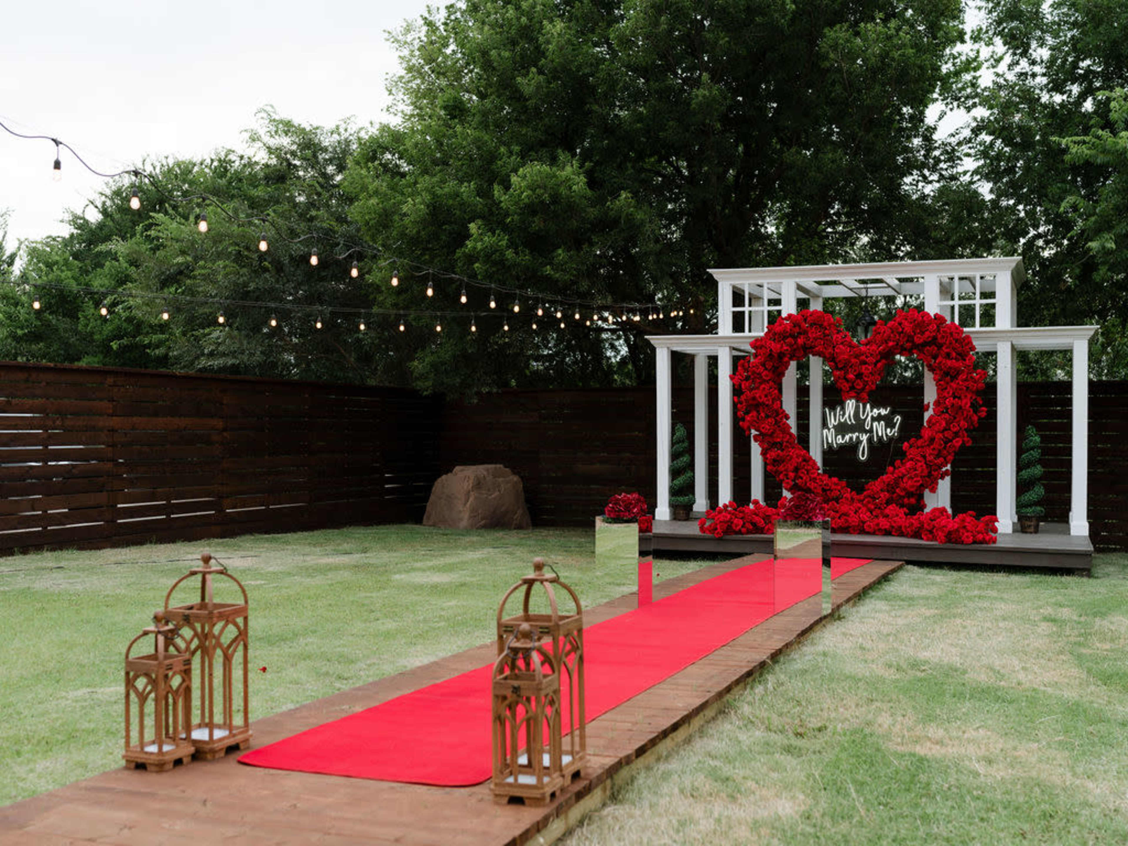 A wedding proposal setup features a large heart made of red roses at the center, flanked by greenery, with a red carpet leading up to it, string lights overhead, and decorative lanterns on either side.