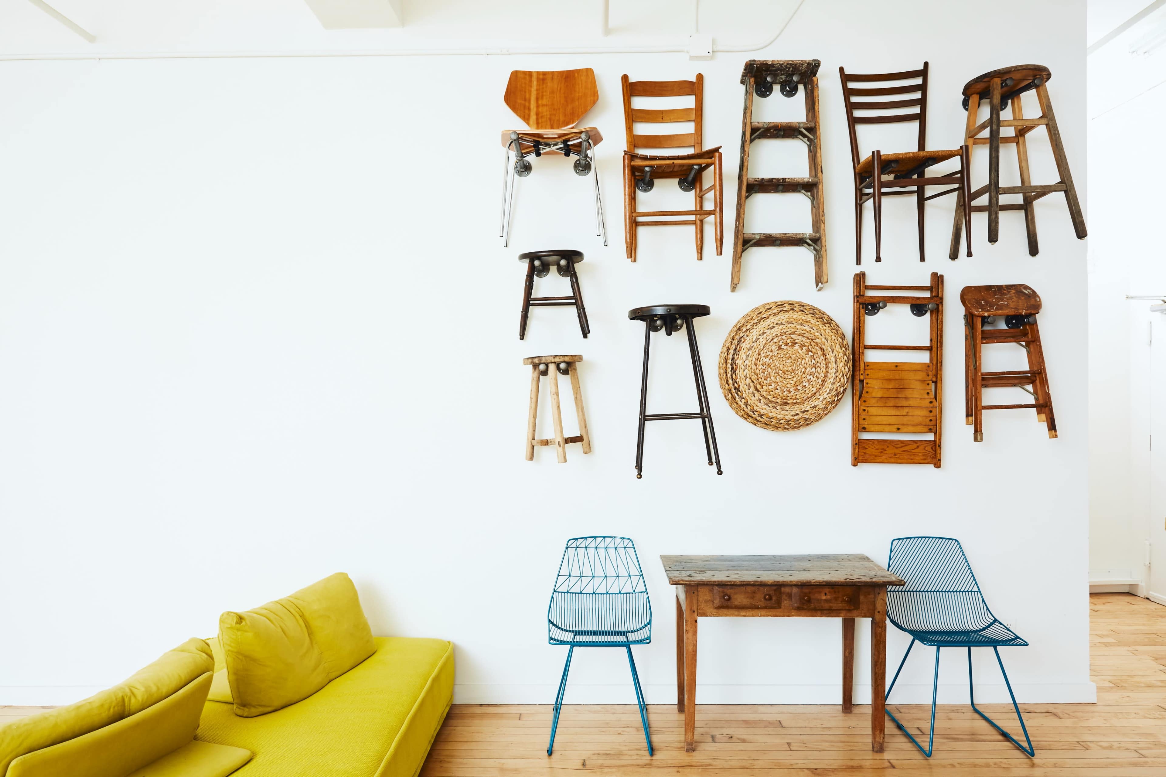 A wall features various wooden chairs arranged in an artistic display above a green couch and a wooden table with two metal chairs.