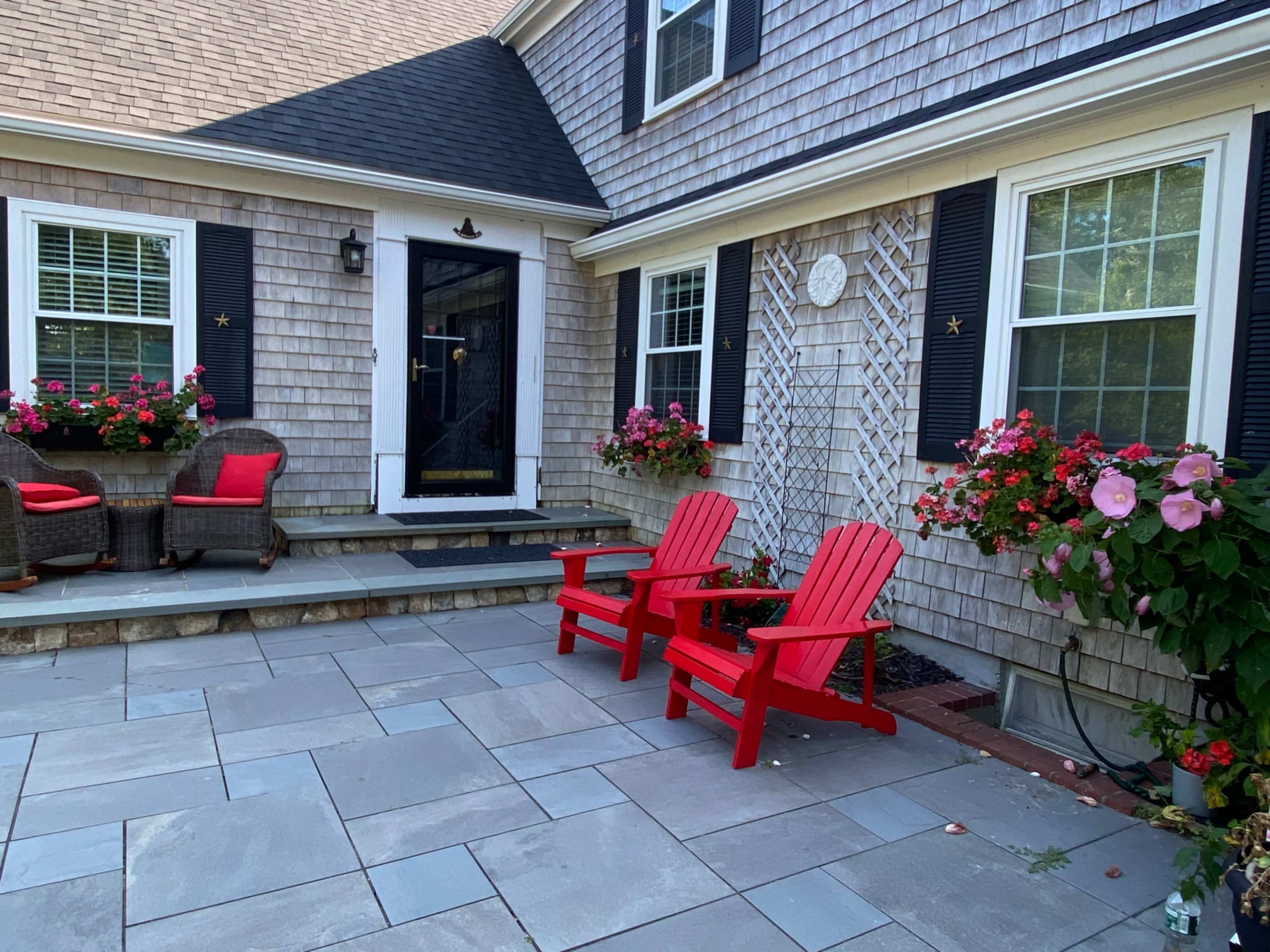 The patio features two red Adirondack chairs and a seating area surrounded by flowering planters and a stone walkway.