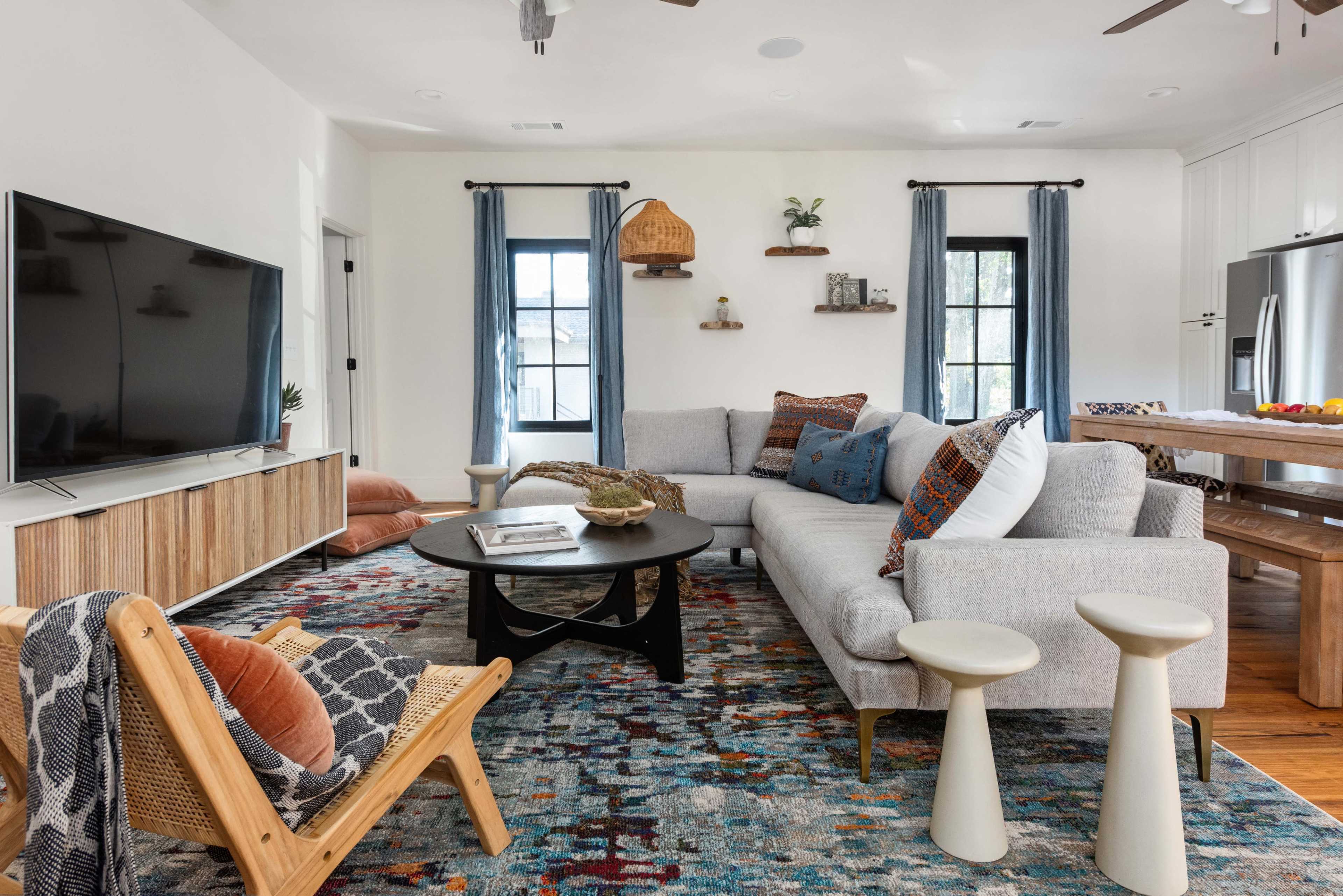 A modern living room features a large gray sofa, a round black coffee table, and a colorful patterned rug, with a TV on a wooden media console and decorative shelves on the wall.