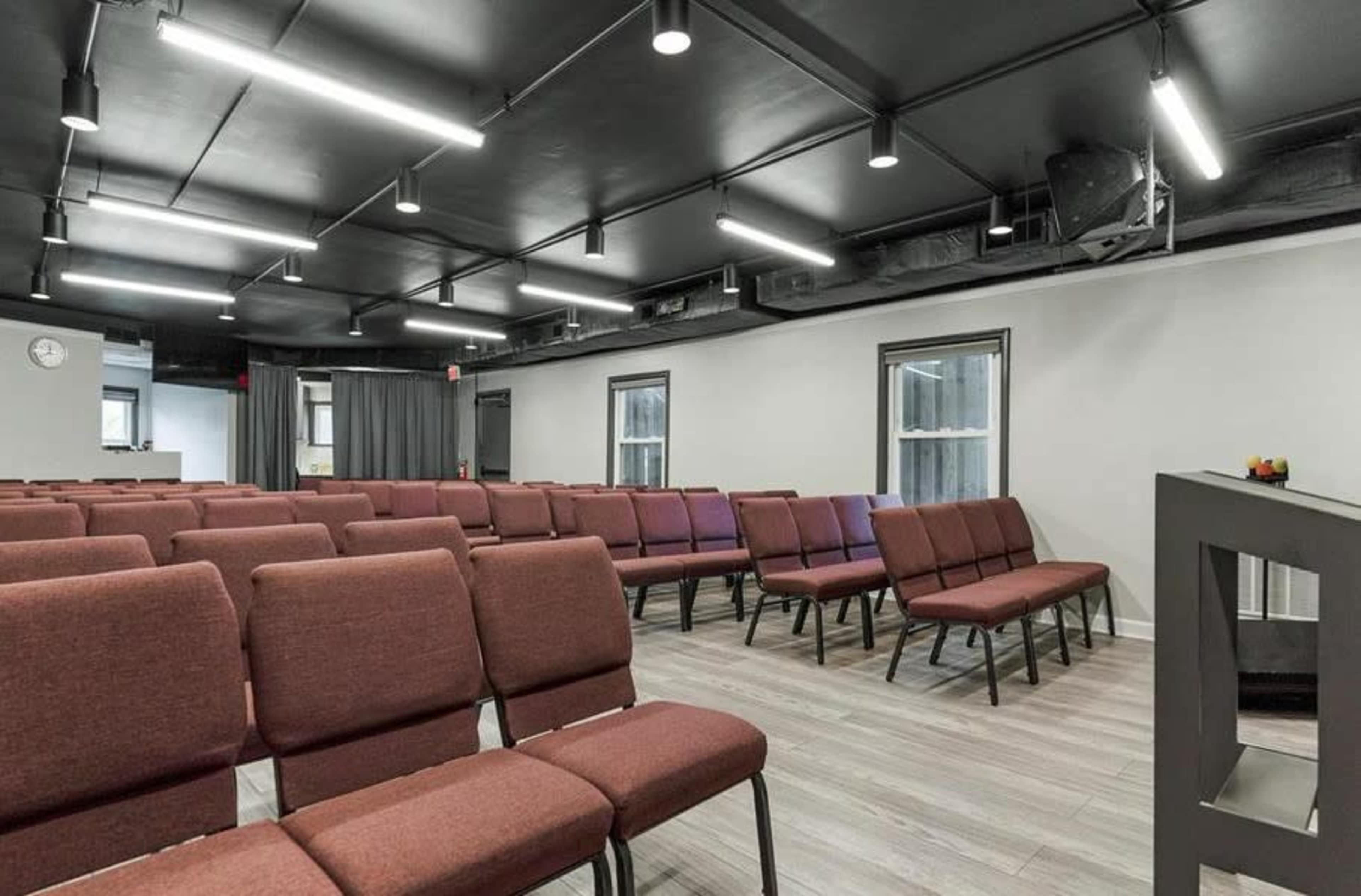 An interior room with multiple rows of maroon chairs arranged for an audience in front of a gray wall with windows.
