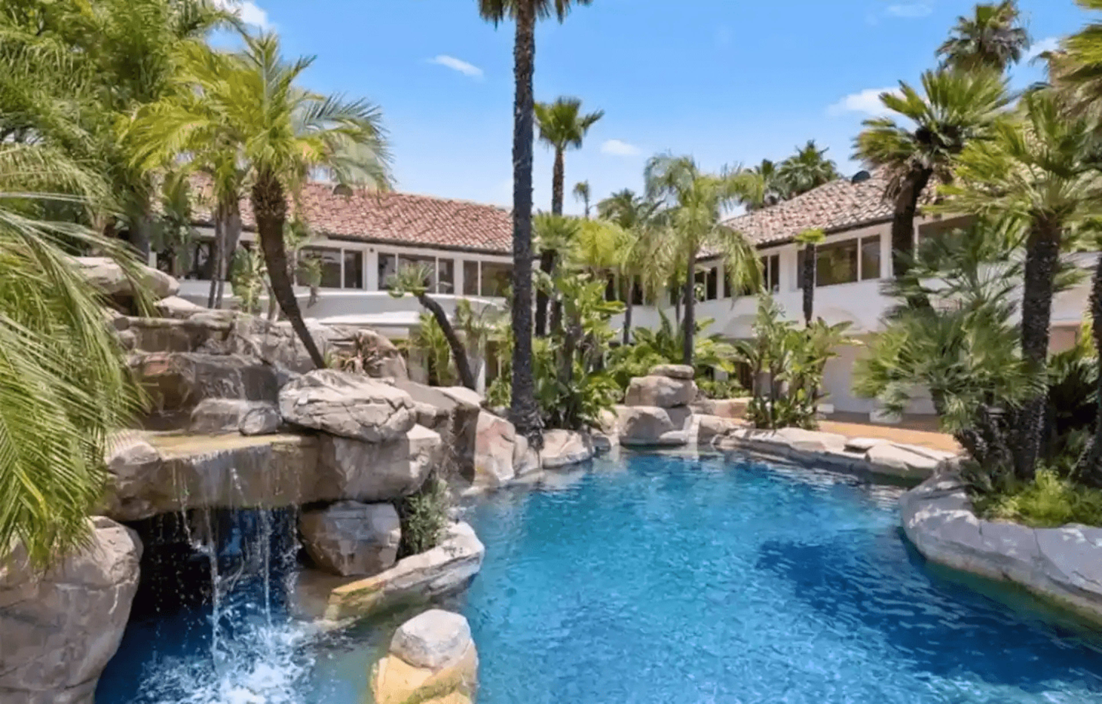 A landscaped backyard with a pool surrounded by palm trees and rock formations, featuring a waterfall.
