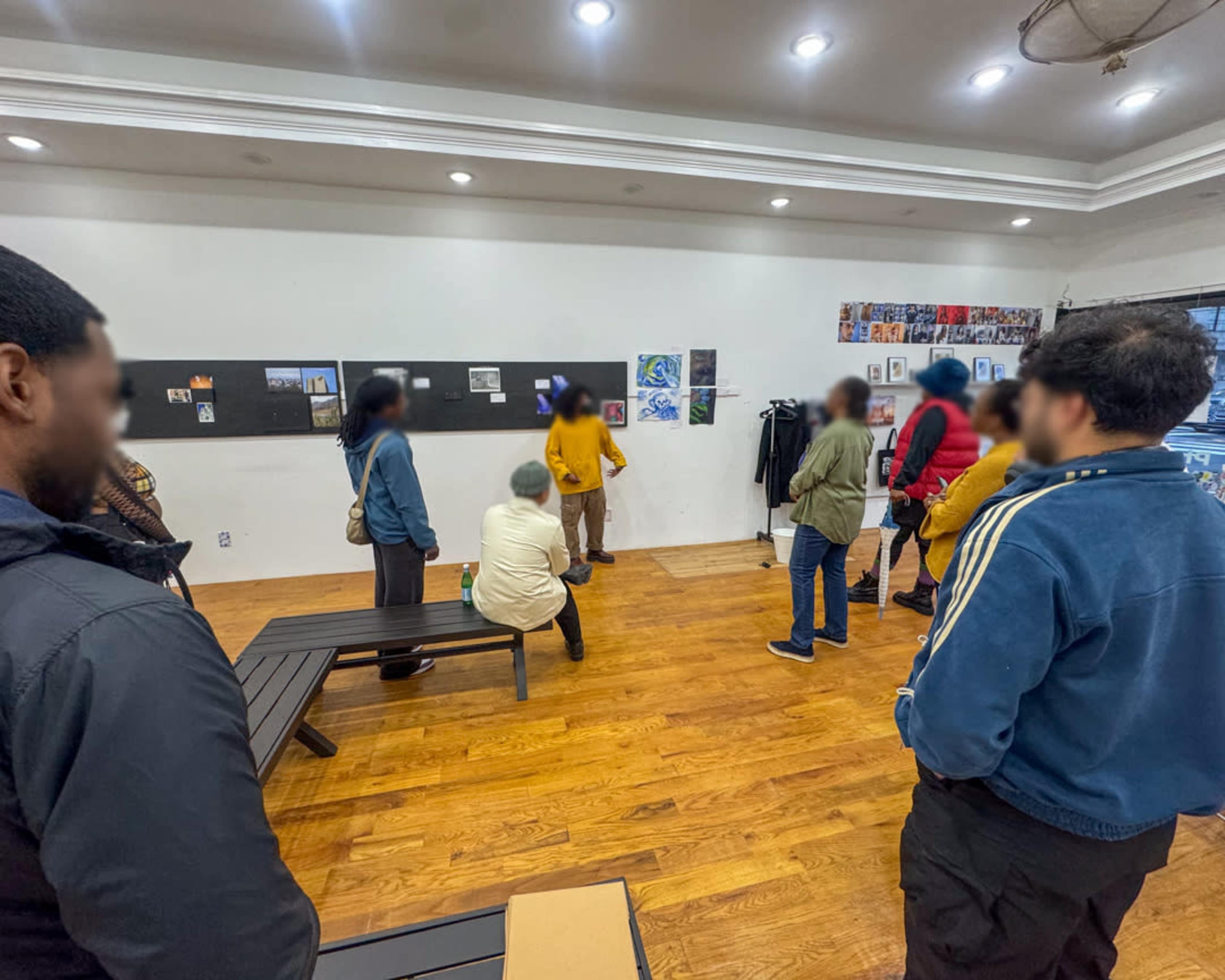 A group of people is gathered in a spacious room with wooden floors, engaging with artwork displayed on a black wall.