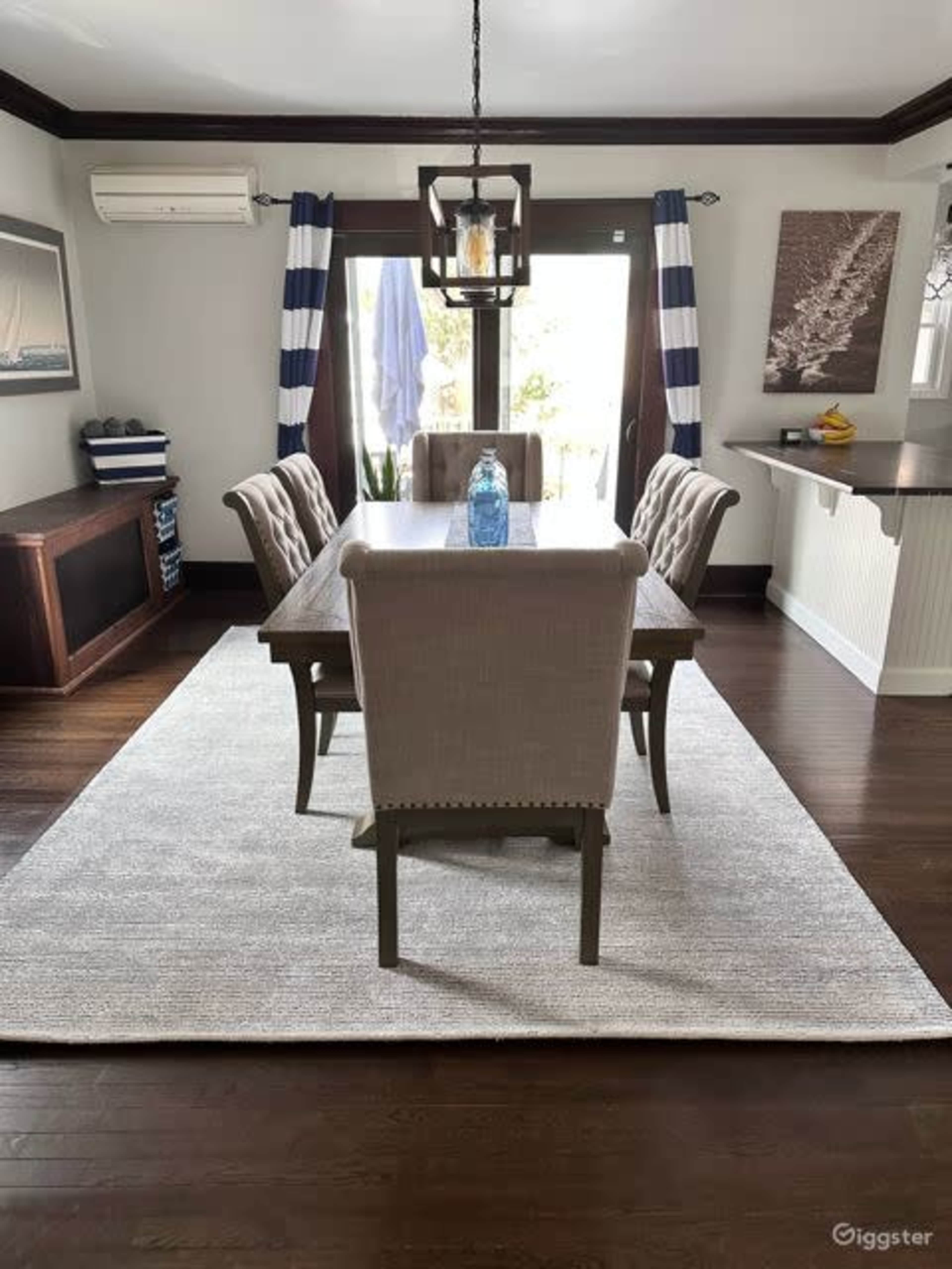 A dining room featuring a rectangular wooden table surrounded by upholstered chairs, with a rug underneath and large windows draped with striped curtains.